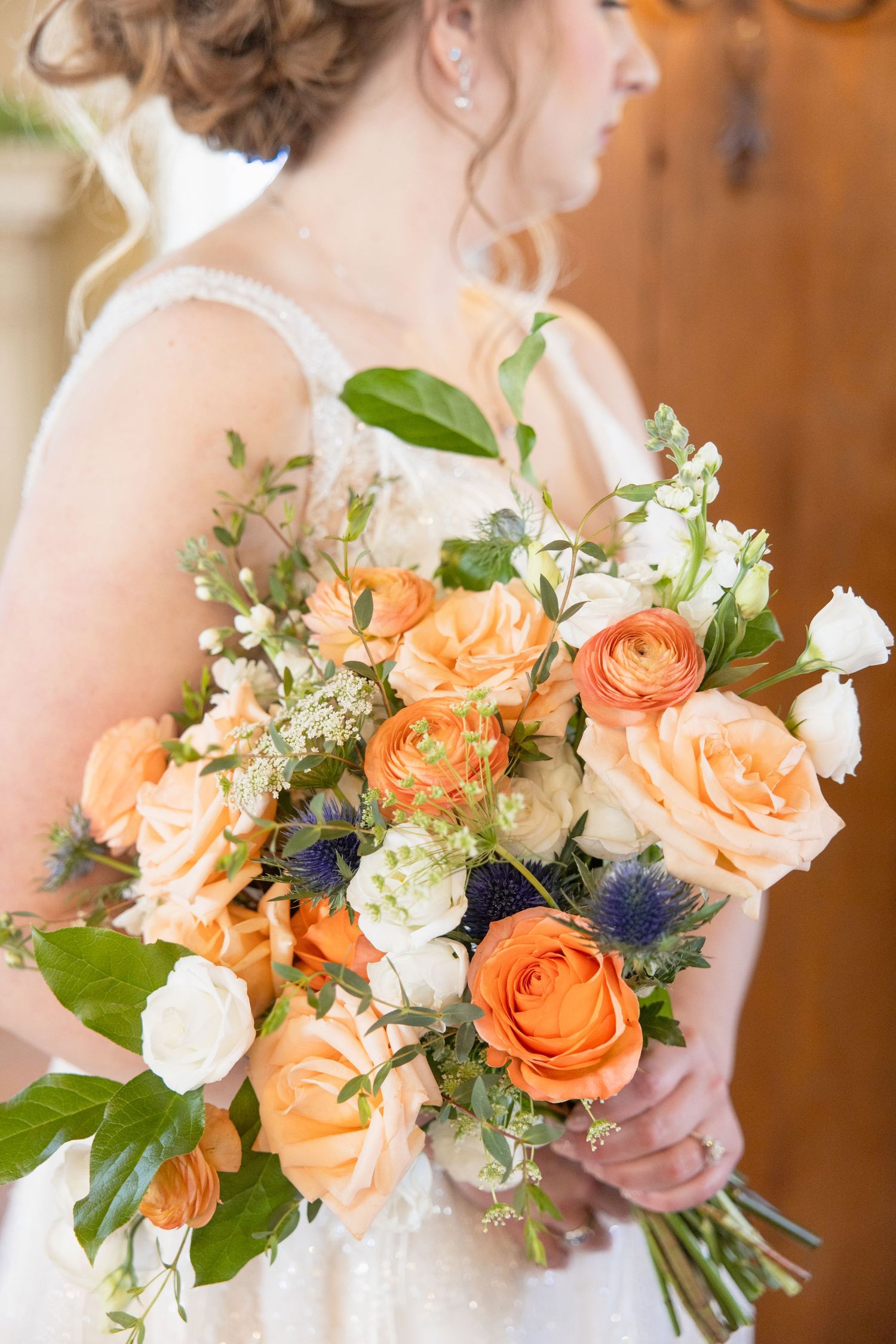 Bride holding a lush wedding bouquet of peach roses, ranunculus, white blooms, and thistle with greenery.