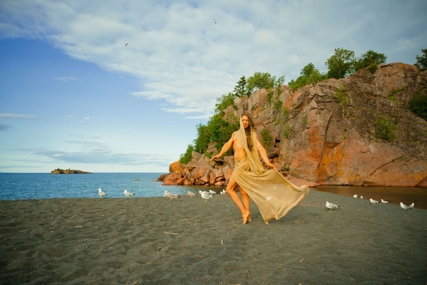 Woman in a flowing golden dress dances on a sandy beach beside rust-colored rocky cliffs, with seagulls and calm blue water behind her.