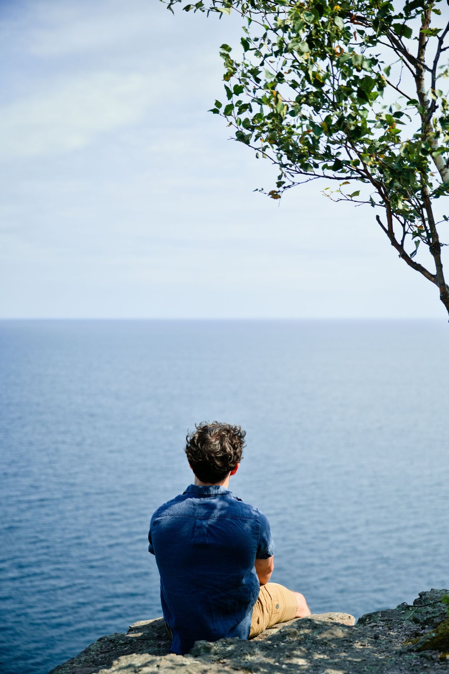 Man sitting on rocky cliff edge overlooking calm blue ocean beside a tree, viewed from behind on a cloudy day.