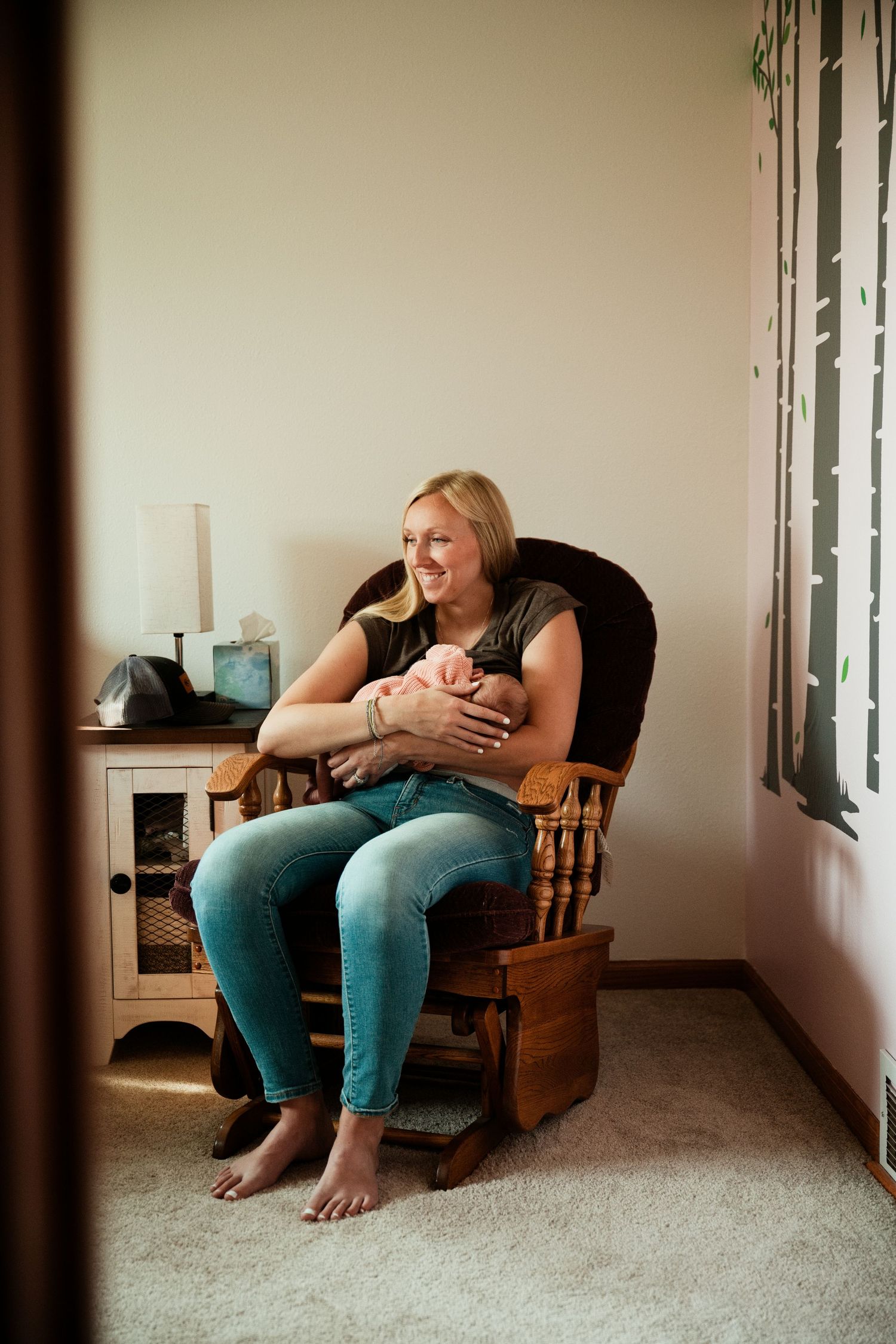 Smiling blonde mother sitting in wooden rocking chair holding newborn baby in cozy nursery room.