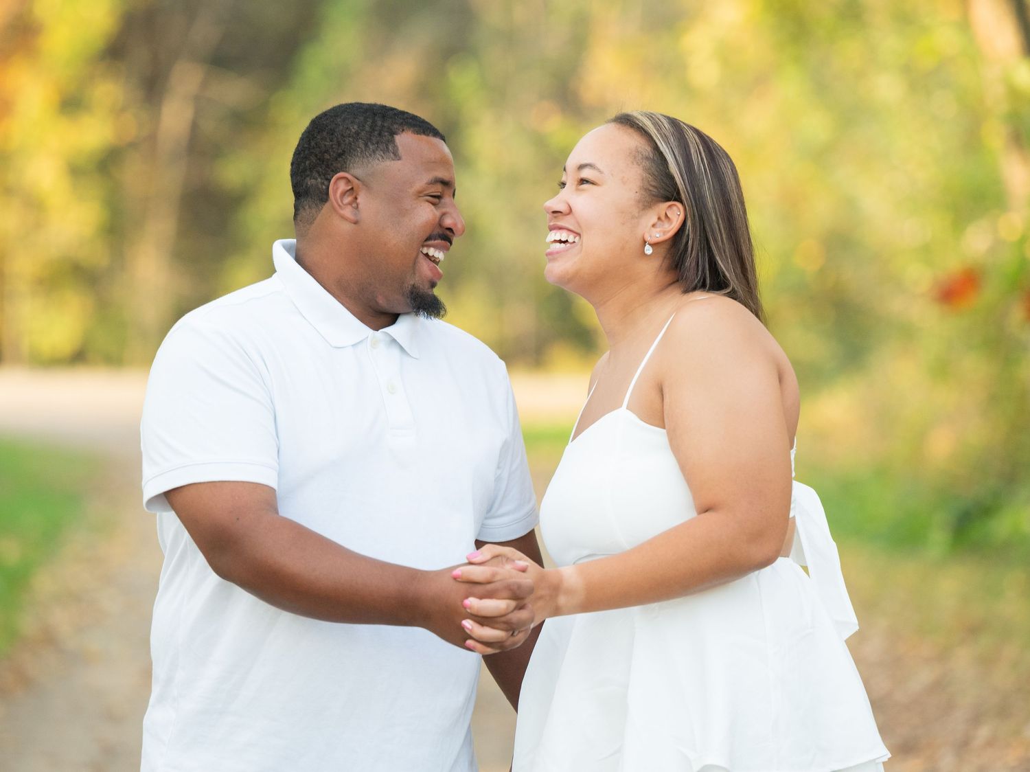 Central Minnesota Best Engagement Photographer Happy couple in white outfits holding hands and laughing together outdoors in a park setting.