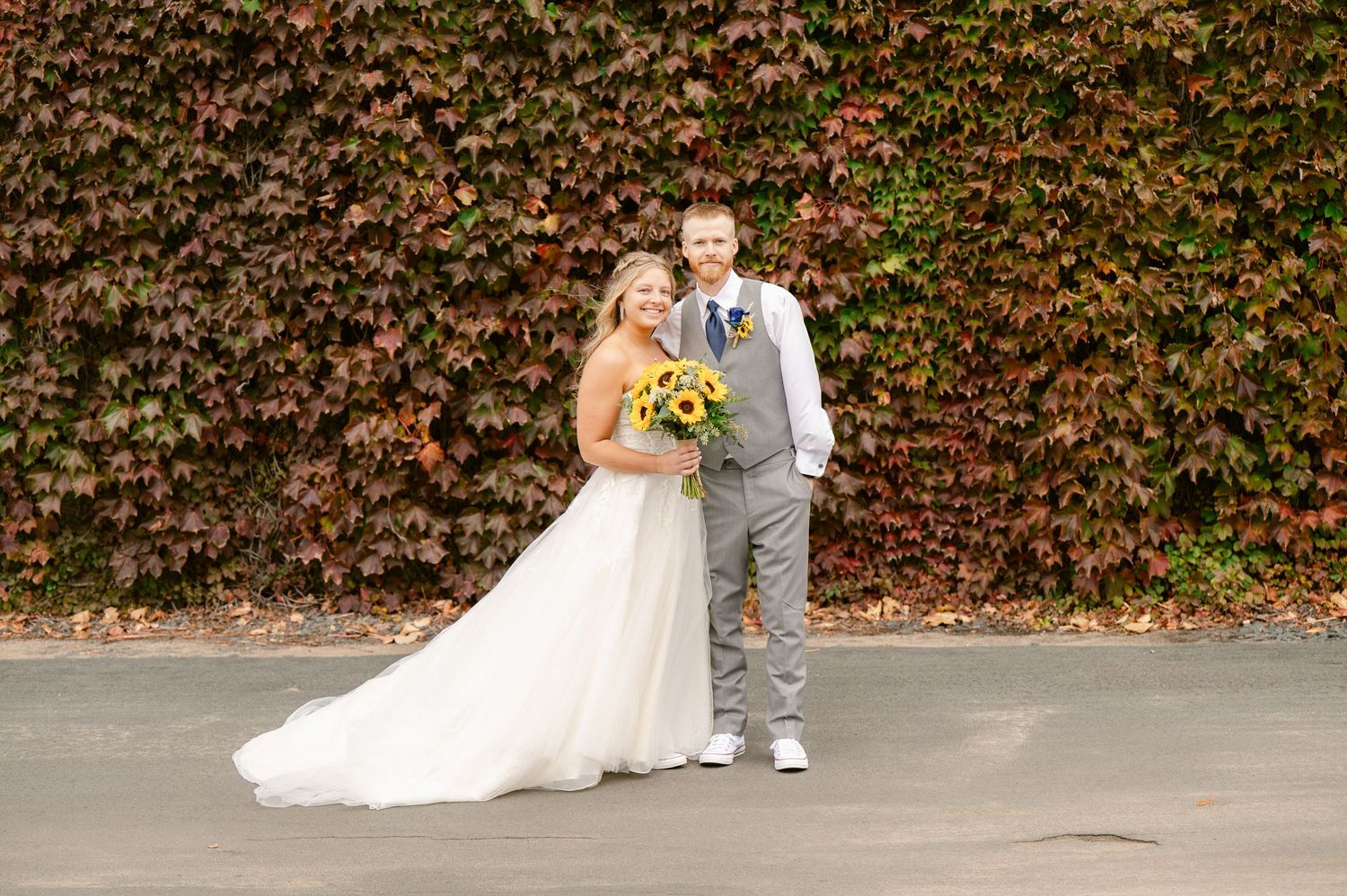 Central MN Wedding Photos | Bride and groom posing with sunflowers before a vibrant red autumn ivy wall in fall wedding portraits.