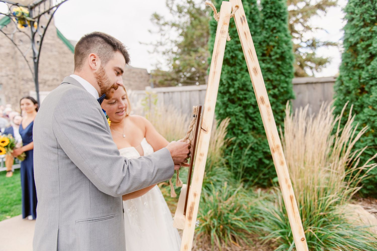 Central MN Wedding Photos - Bride and groom sign wedding document together at rustic wooden easel during outdoor ceremony.