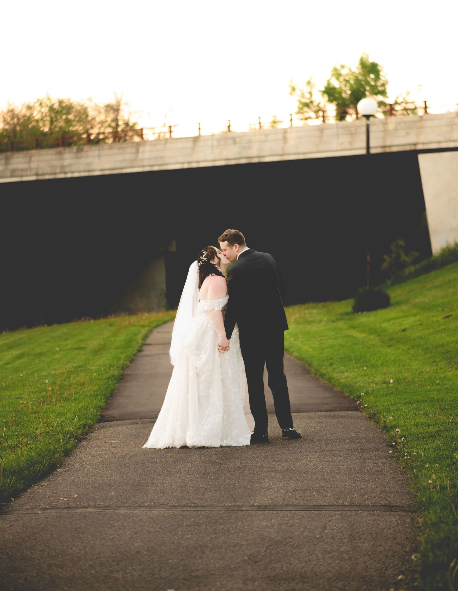 Central MN Wedding Photos - Bride and groom sharing a kiss on a pathway surrounded by lush green grass at golden hour during their wedding.