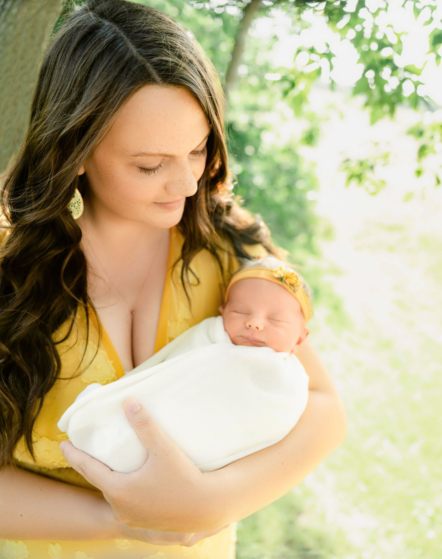 St Cloud newborn Photos of A mother in a yellow dress tenderly holds her swaddled newborn baby outdoors among green trees.
