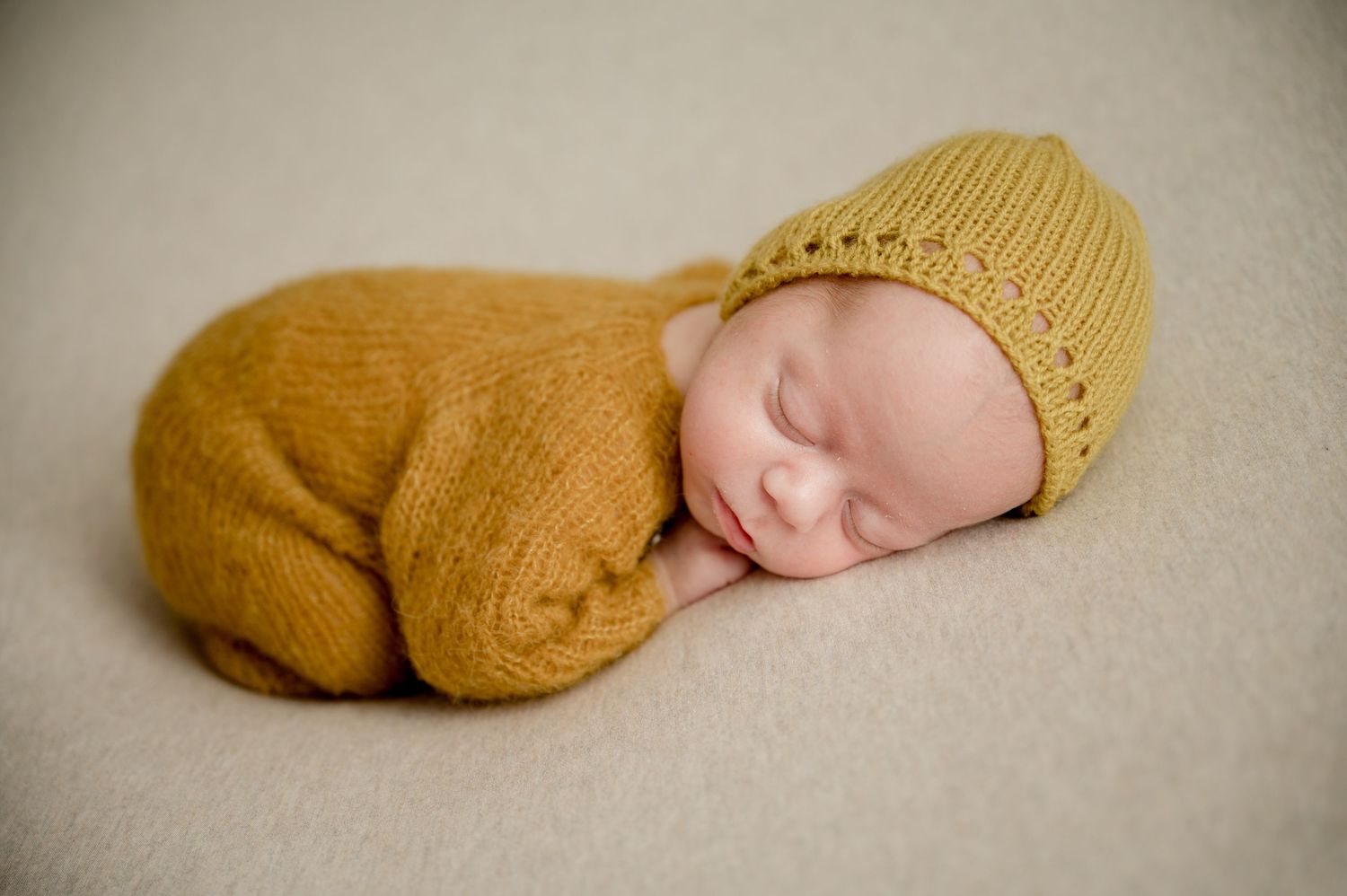 Sleeping newborn baby in mustard yellow knit outfit and bonnet on cream blanket.