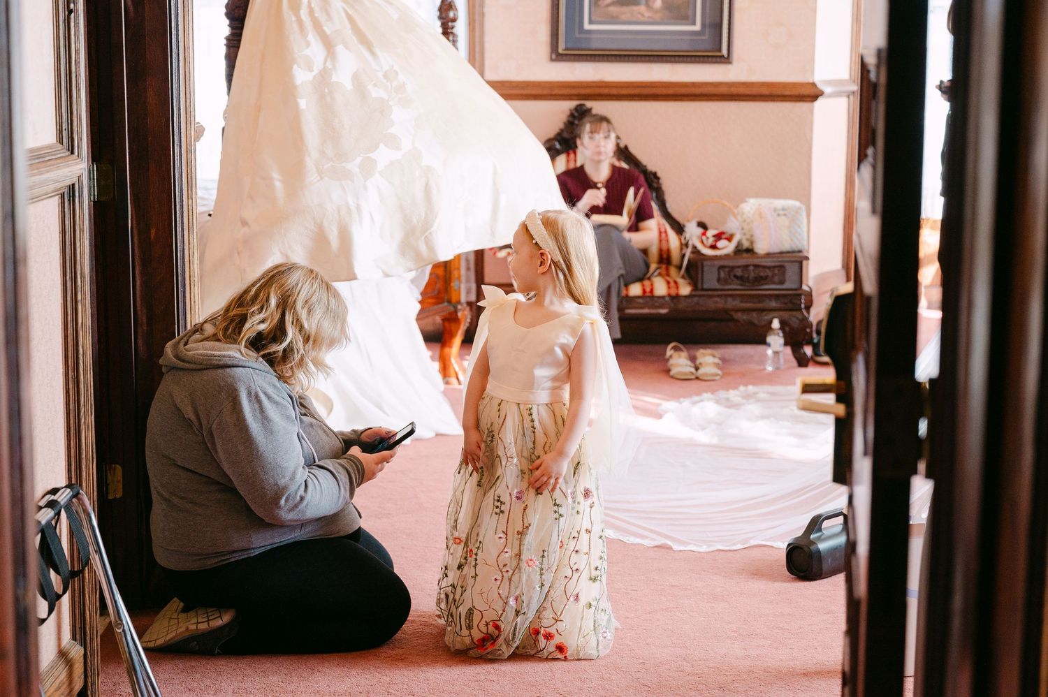 Central MN Wedding Photos | Woman helping young flower girl in white dress with veil during wedding preparation indoors.