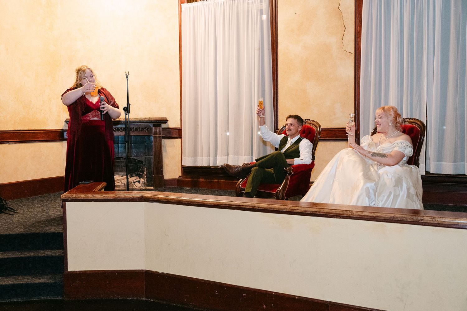 Central MN Wedding Photos | Bride and groom seated in ornate chairs on stage while guest in burgundy reads during wedding reception.