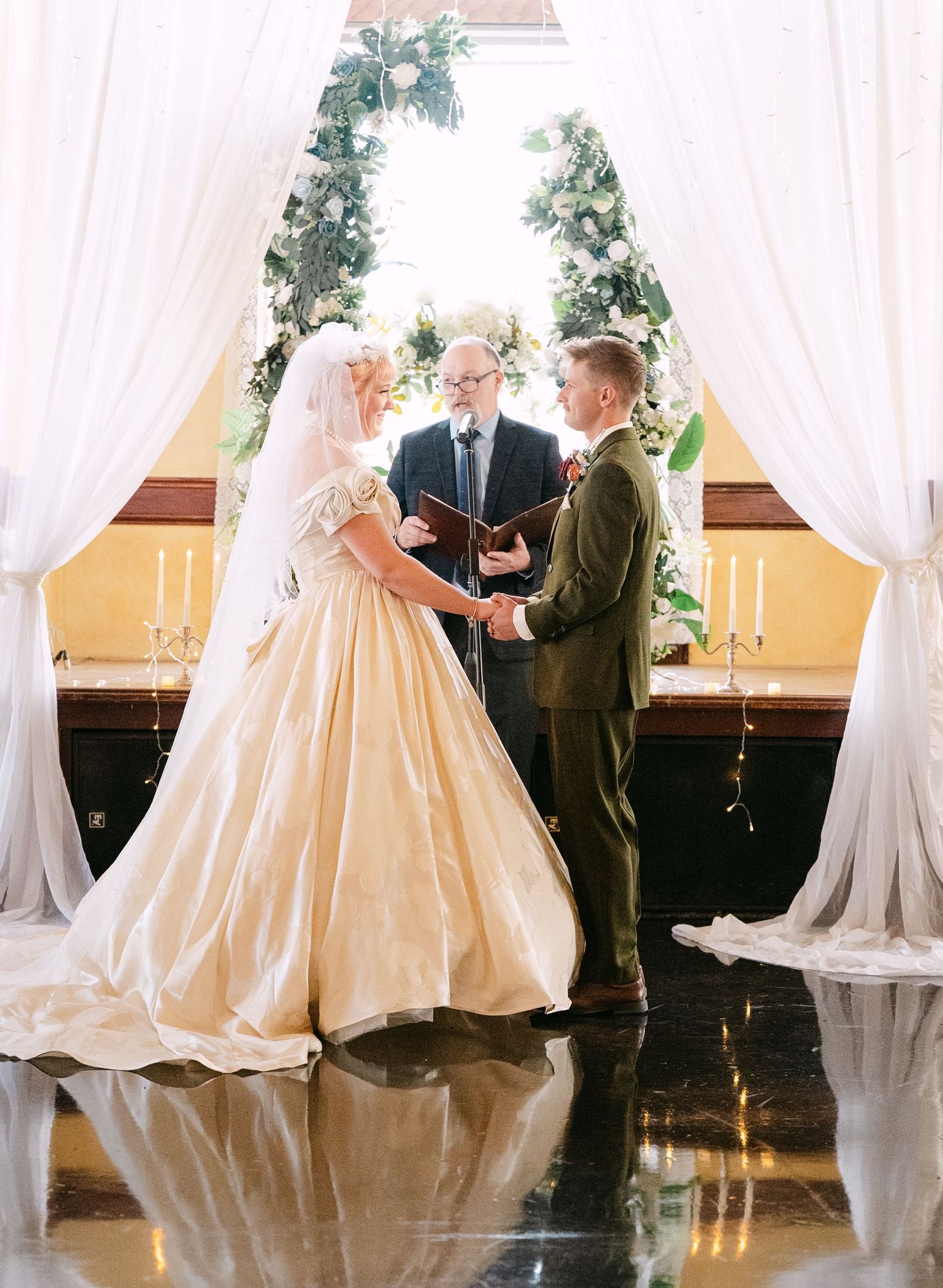 Central MN Wedding Photos | Bride and groom exchanging vows at floral arch altar with officiant during elegant wedding ceremony.