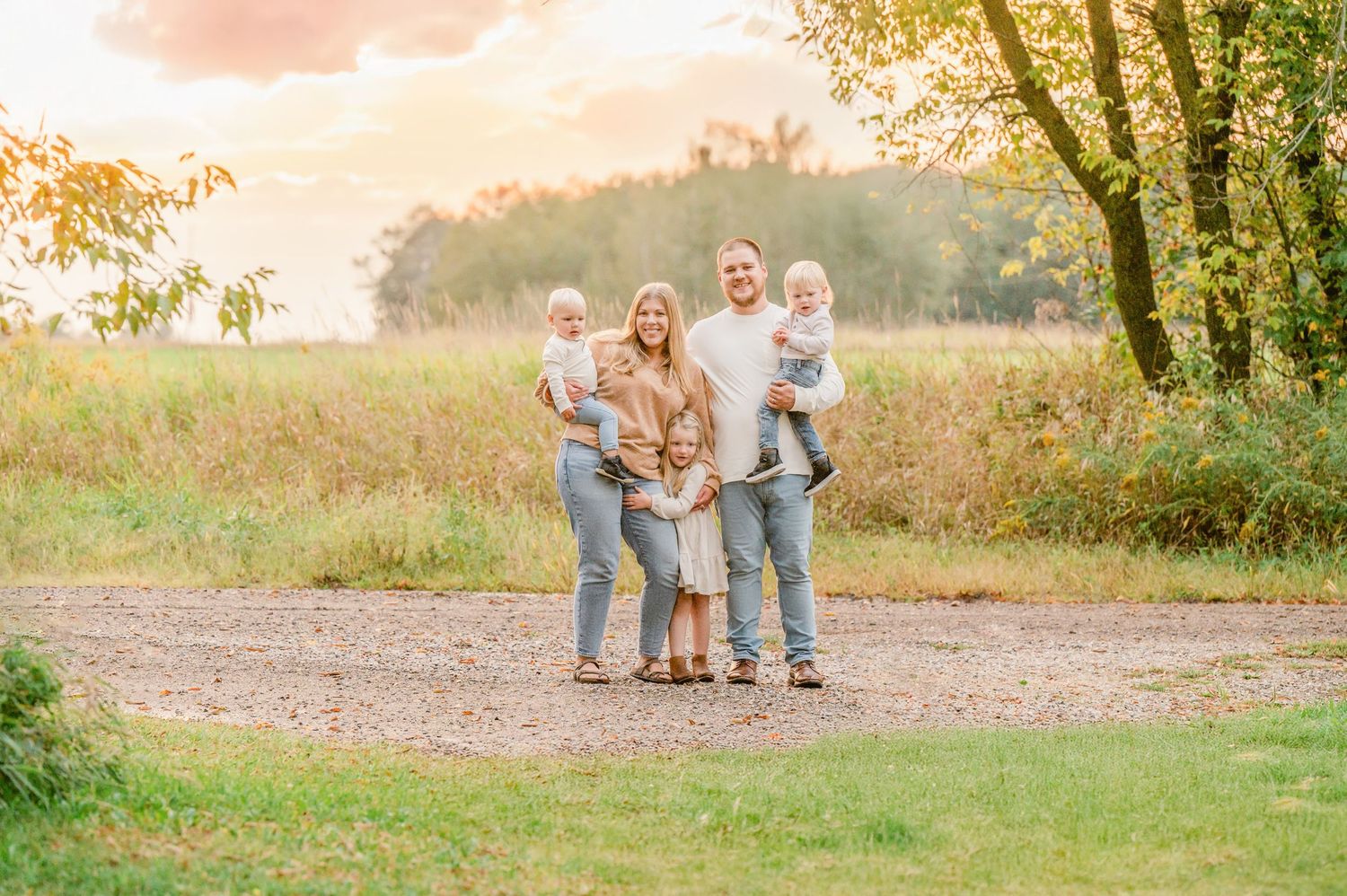 A family enjoys a sunset photoshoot on a dirt path surrounded by golden field grass and trees.