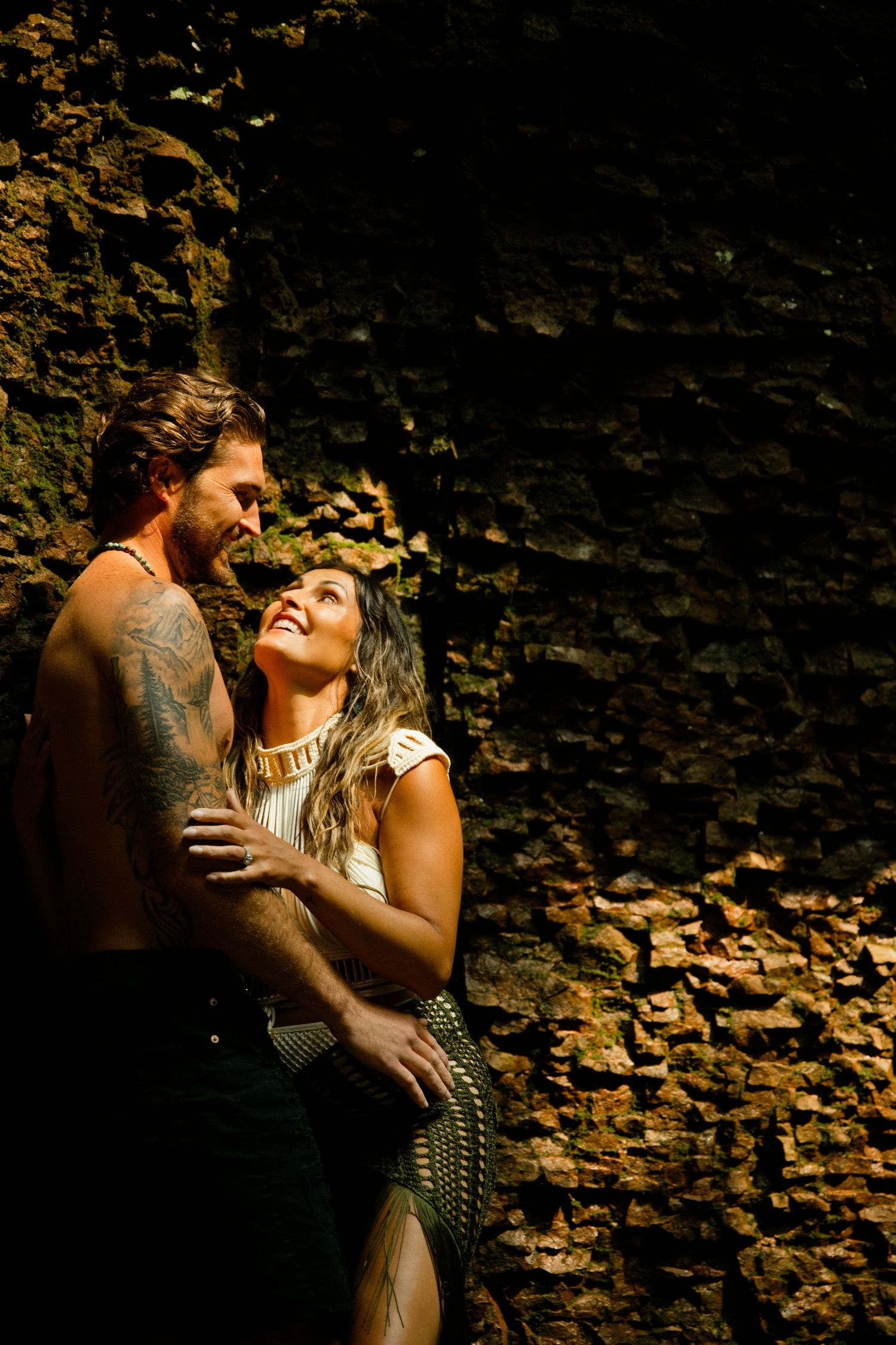 A shirtless tattooed man embraces a smiling woman against a rustic stone wall in a romantic outdoor setting.