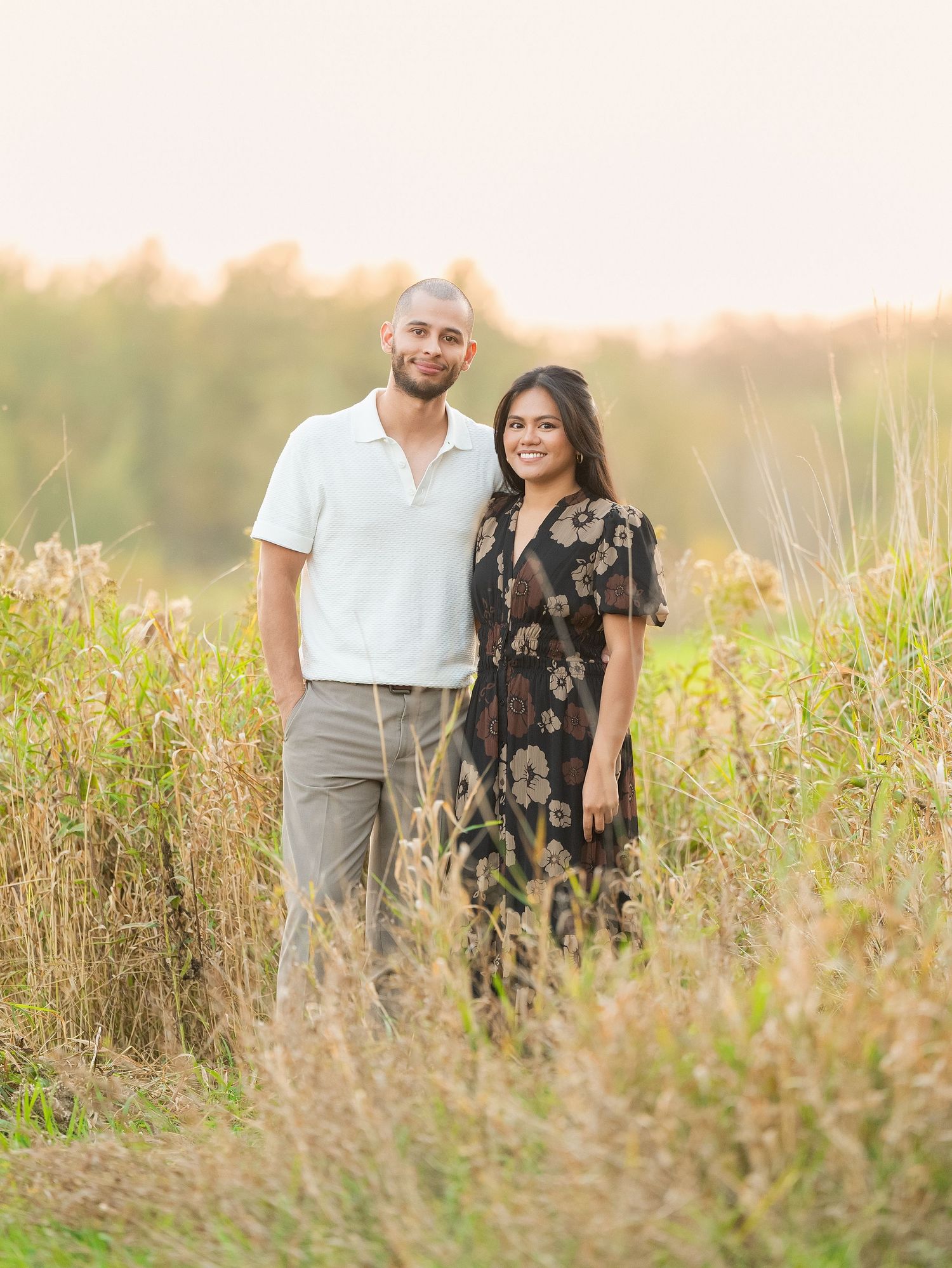 A couple stands together in a golden field at sunset while posing for romantic outdoor portraits.