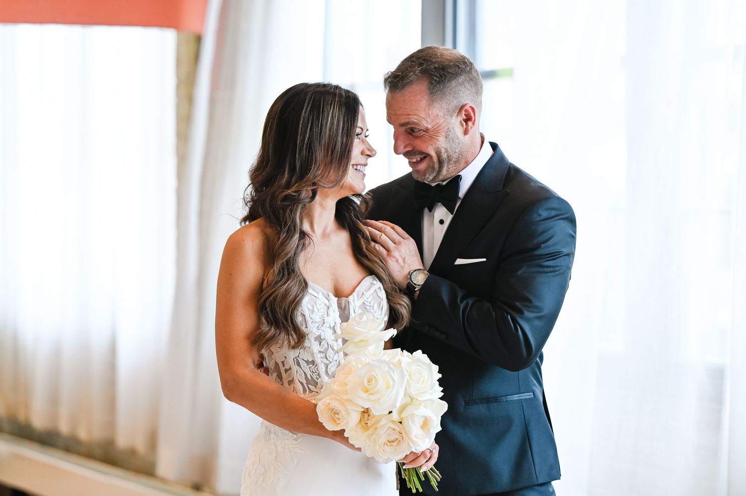 Tender embrace between newlyweds in front of white drapes and natural lighting.