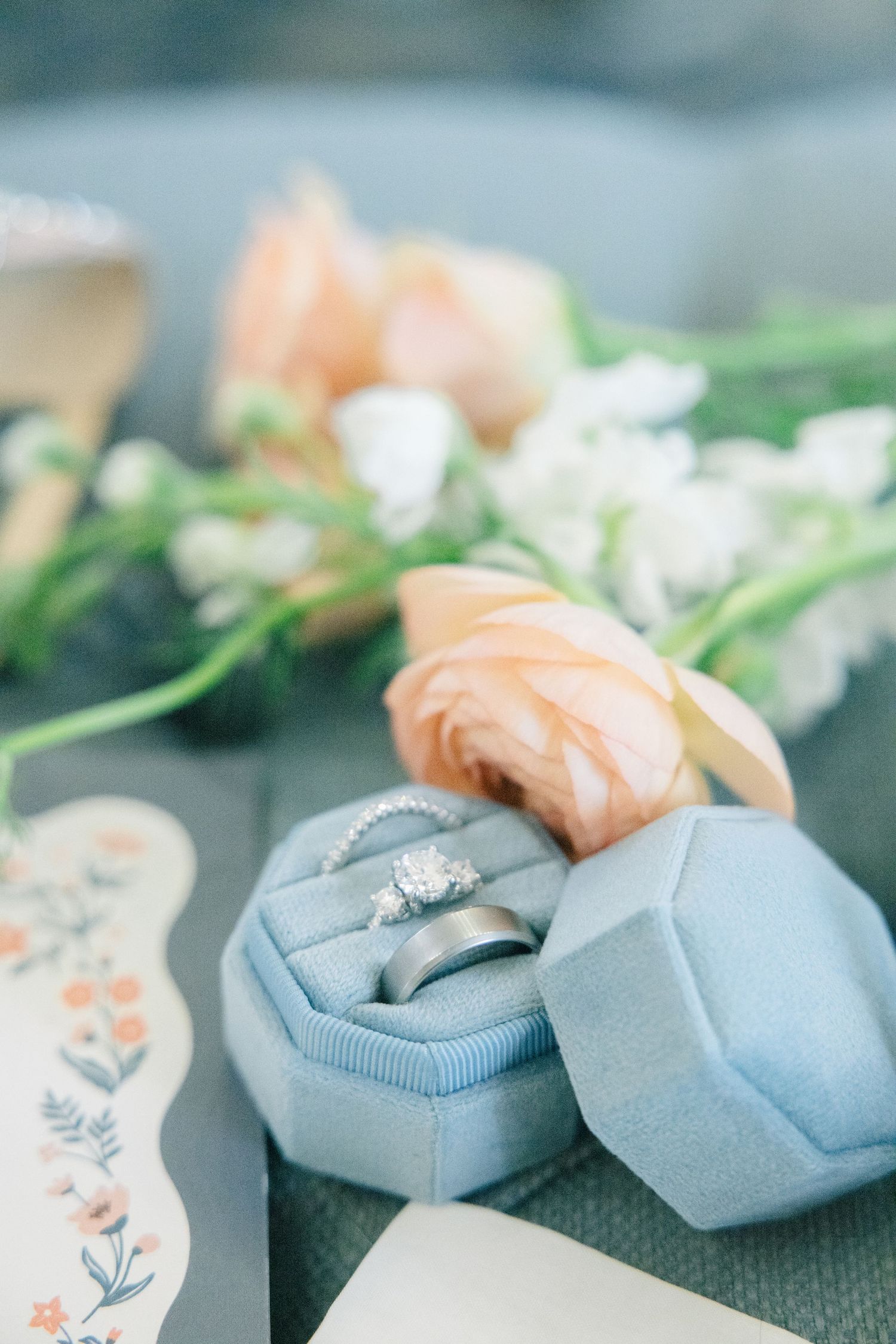 Blue velvet hexagonal ring box holding diamond engagement ring and wedding band beside peach roses.