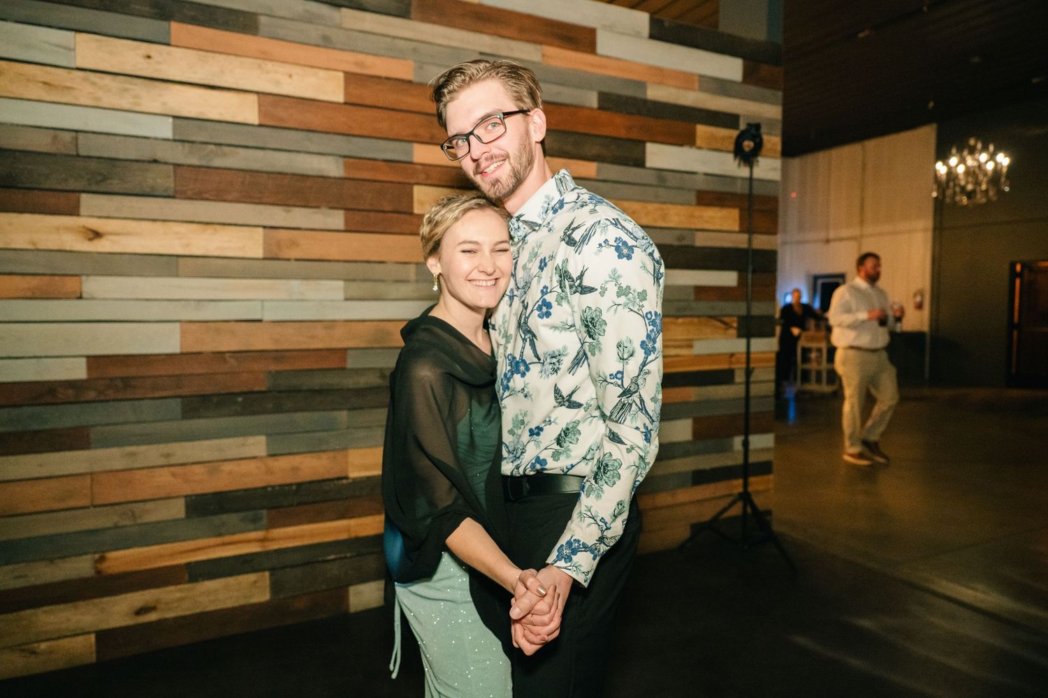 Couple poses holding hands in front of rustic reclaimed wood wall at wedding venue.