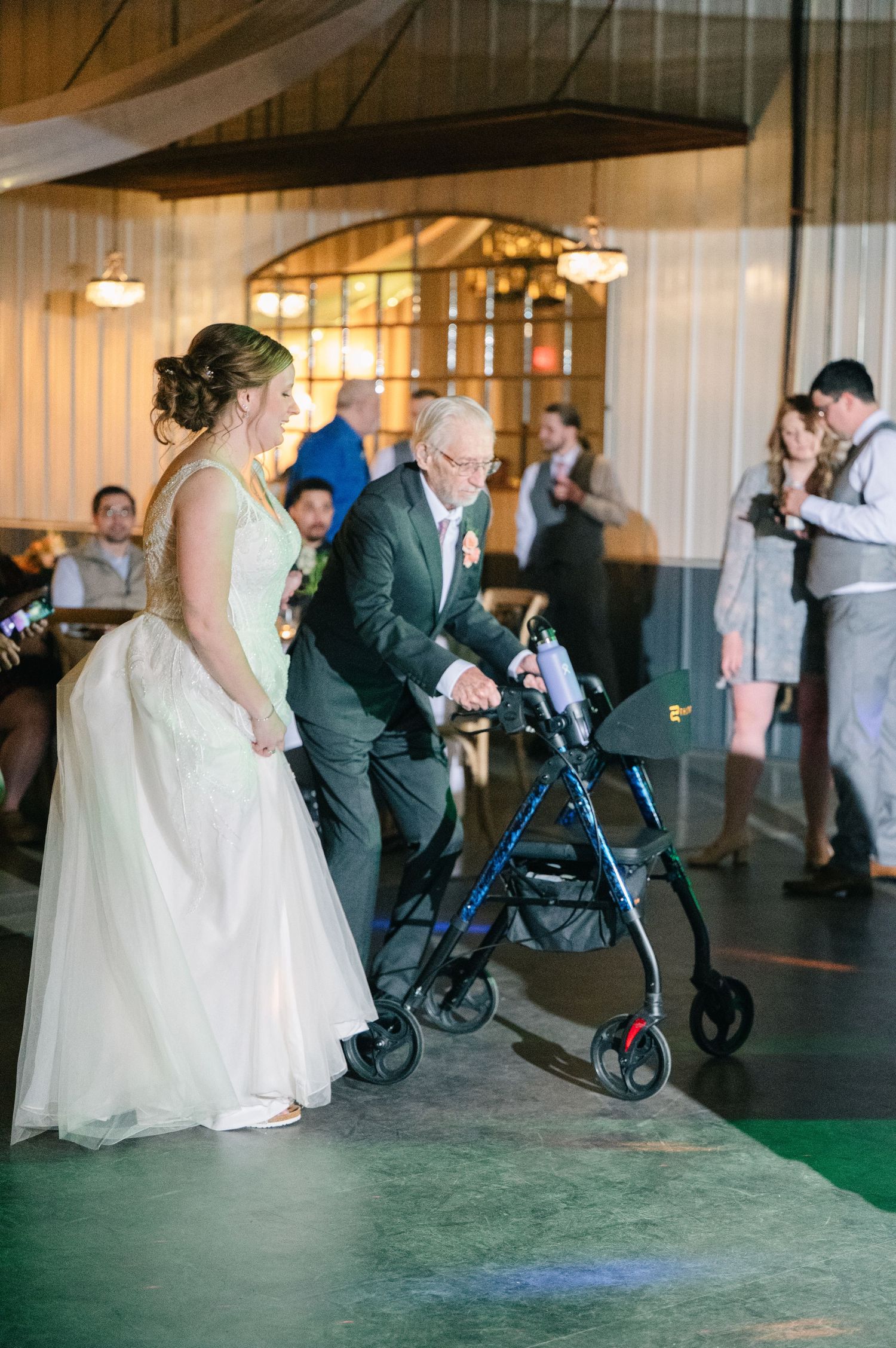 Bride assists elderly grandfather using a walker onto the dance floor at wedding reception.