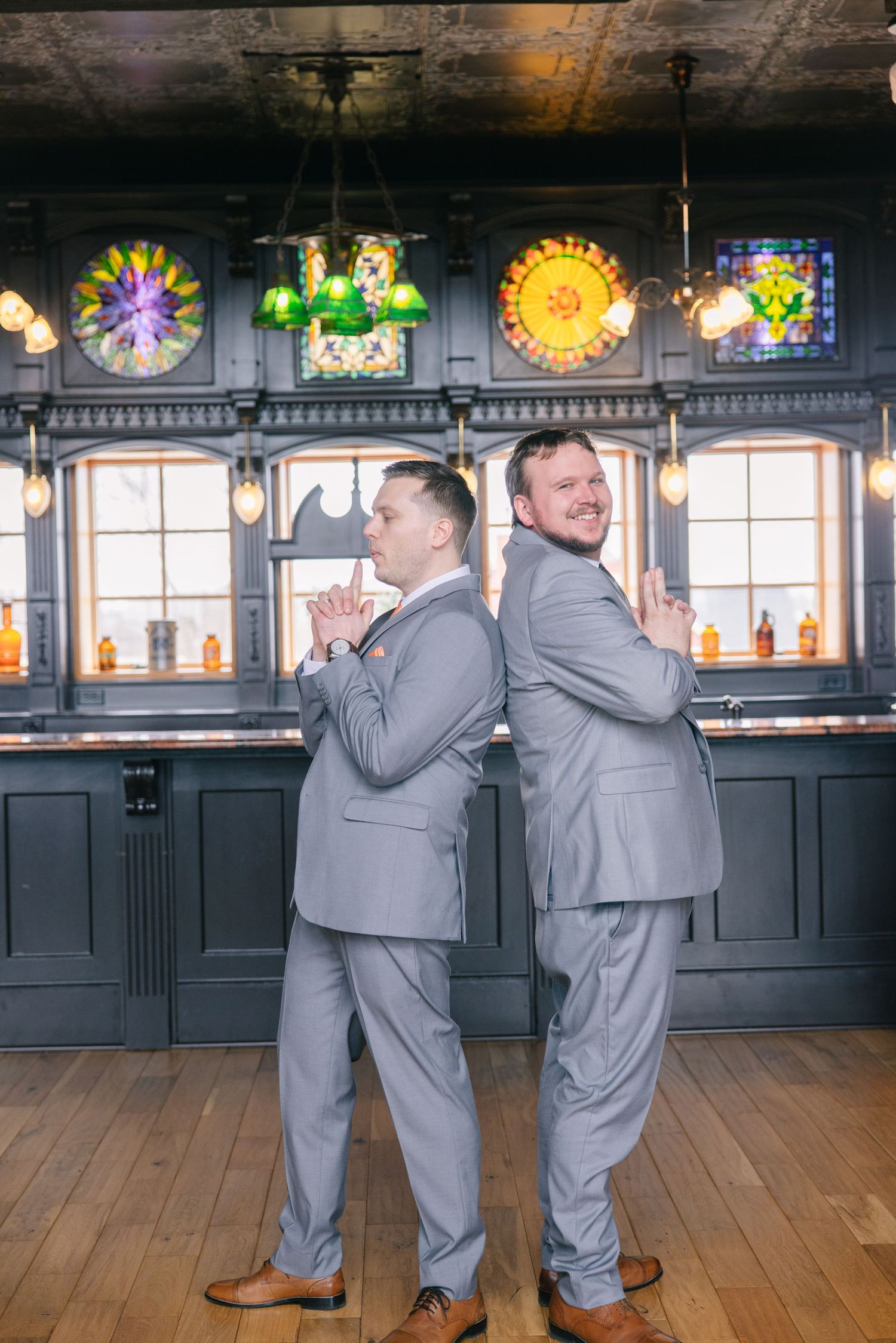 Two grooms in matching gray suits pose back-to-back in a bar with colorful stained glass windows.