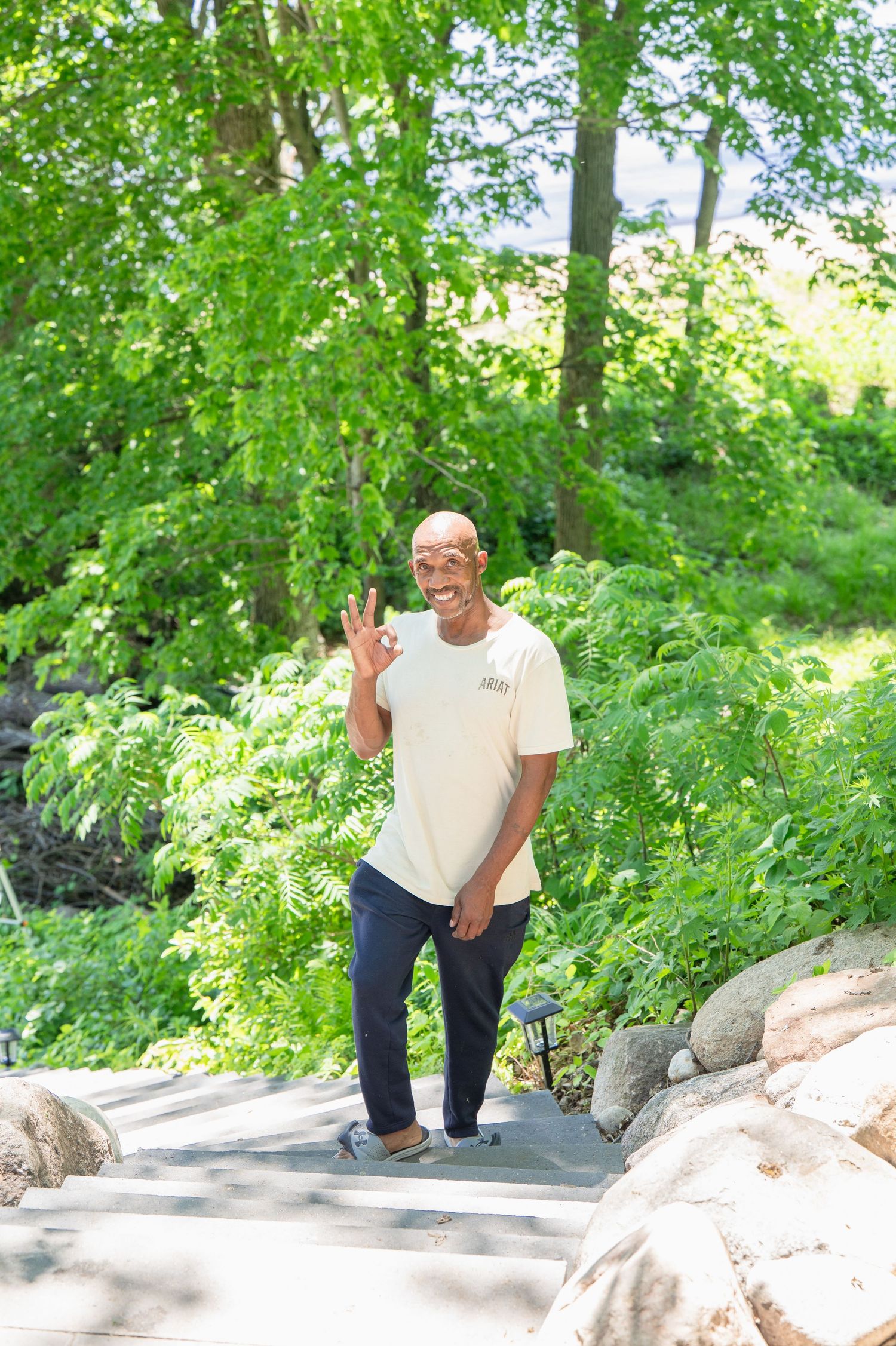 A smiling bald Black man makes an OK gesture on a sunny garden path surrounded by lush green trees and rocks.