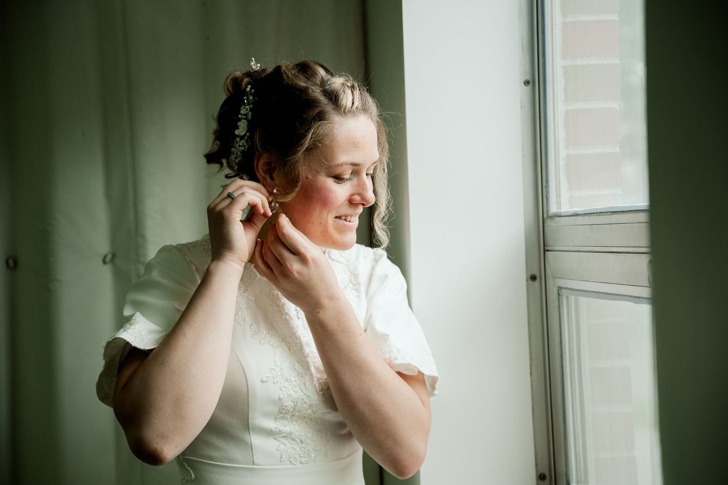 A bride in a white wedding dress adjusts her earrings while standing near a window in soft natural light.