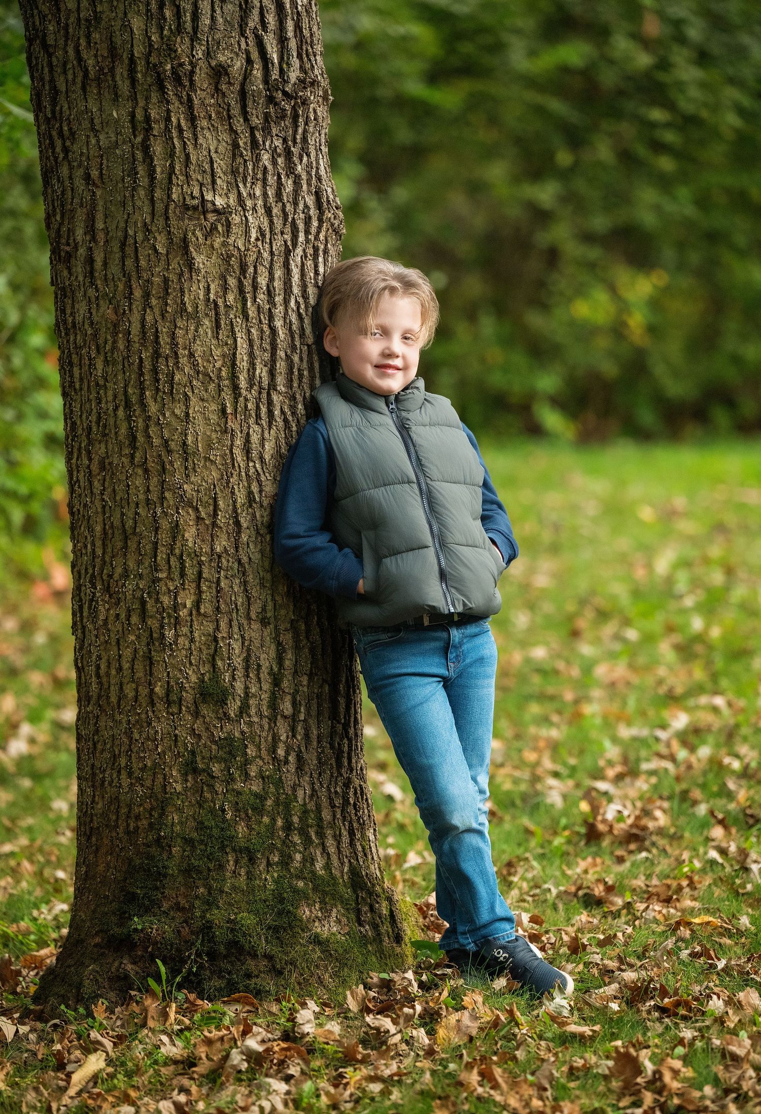 A child in a puffy vest and jeans plays peek-a-boo behind a large tree trunk in an autumn forest setting.