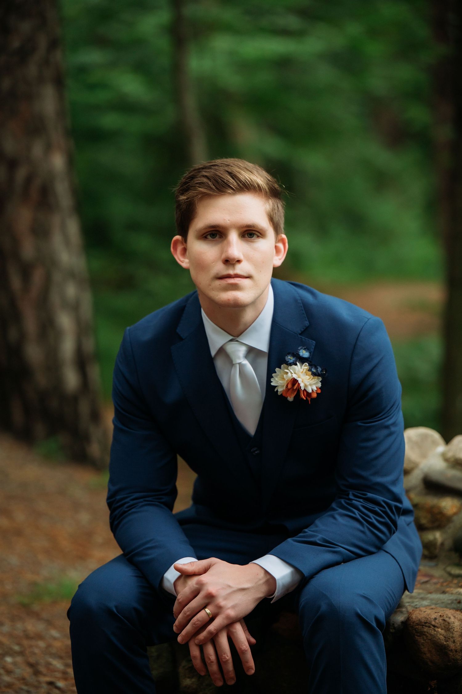 A navy blue suited figure sits contemplatively on natural stone in a wooded setting.