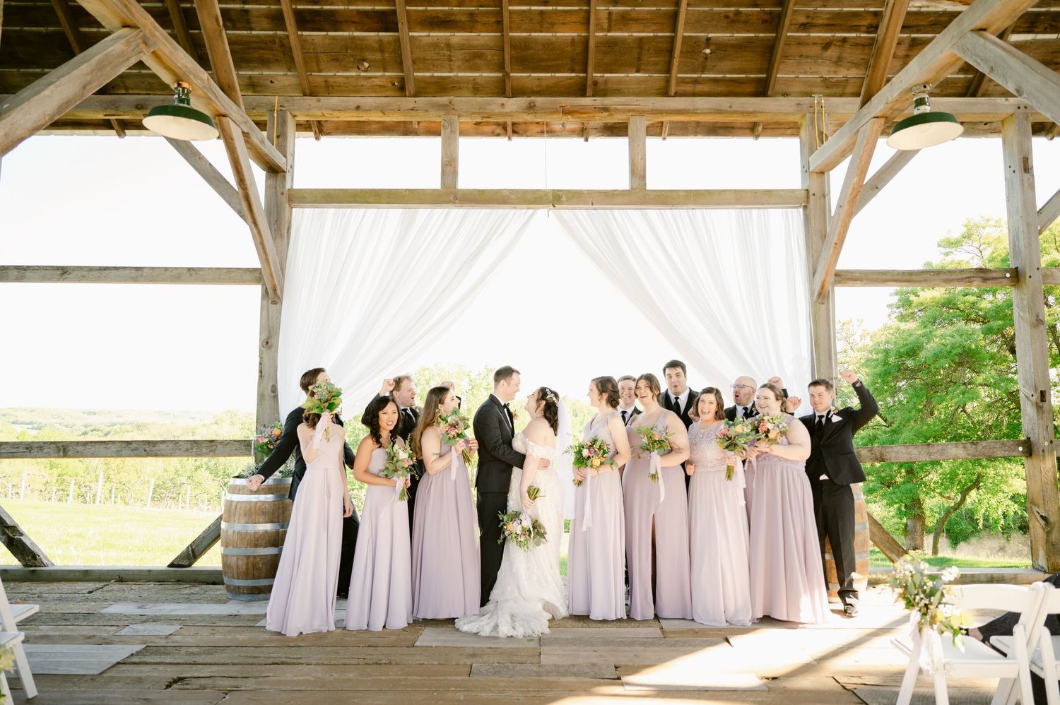 Wedding party poses under rustic wooden pavilion with flowing white curtains and pink bridesmaid dresses.