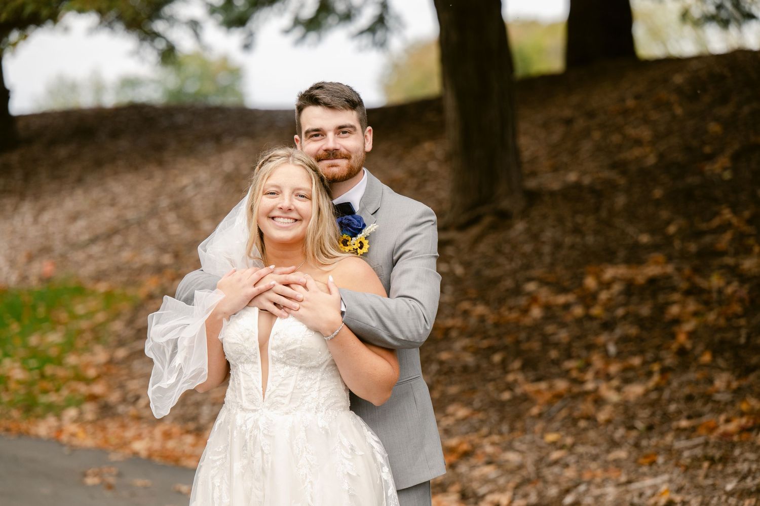 Central MN Wedding Photos | Smiling bride in lace gown with groom embracing her from behind on a tree-lined path.