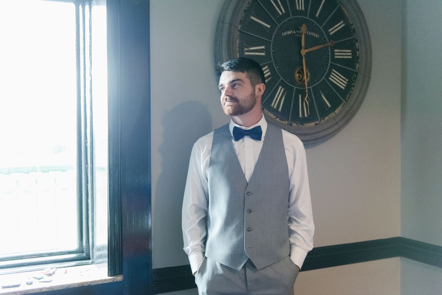 Central MN Wedding Photos | Groom in gray vest and navy bow tie standing by window with vintage clock.