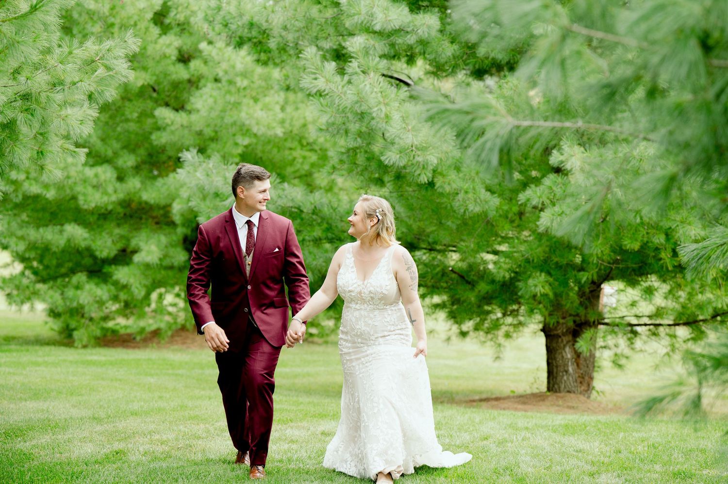 Central MN Wedding Photos - Bride and groom holding hands walking through lush green trees, groom in burgundy suit and bride in lace gown.