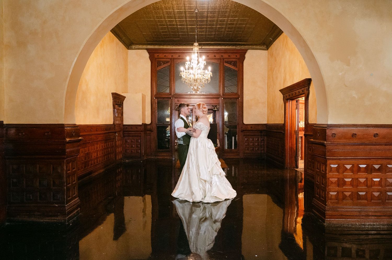Central MN Wedding Photos | Bride and groom share romantic kiss under elegant arch in grand ballroom with chandelier and dark wood paneling.