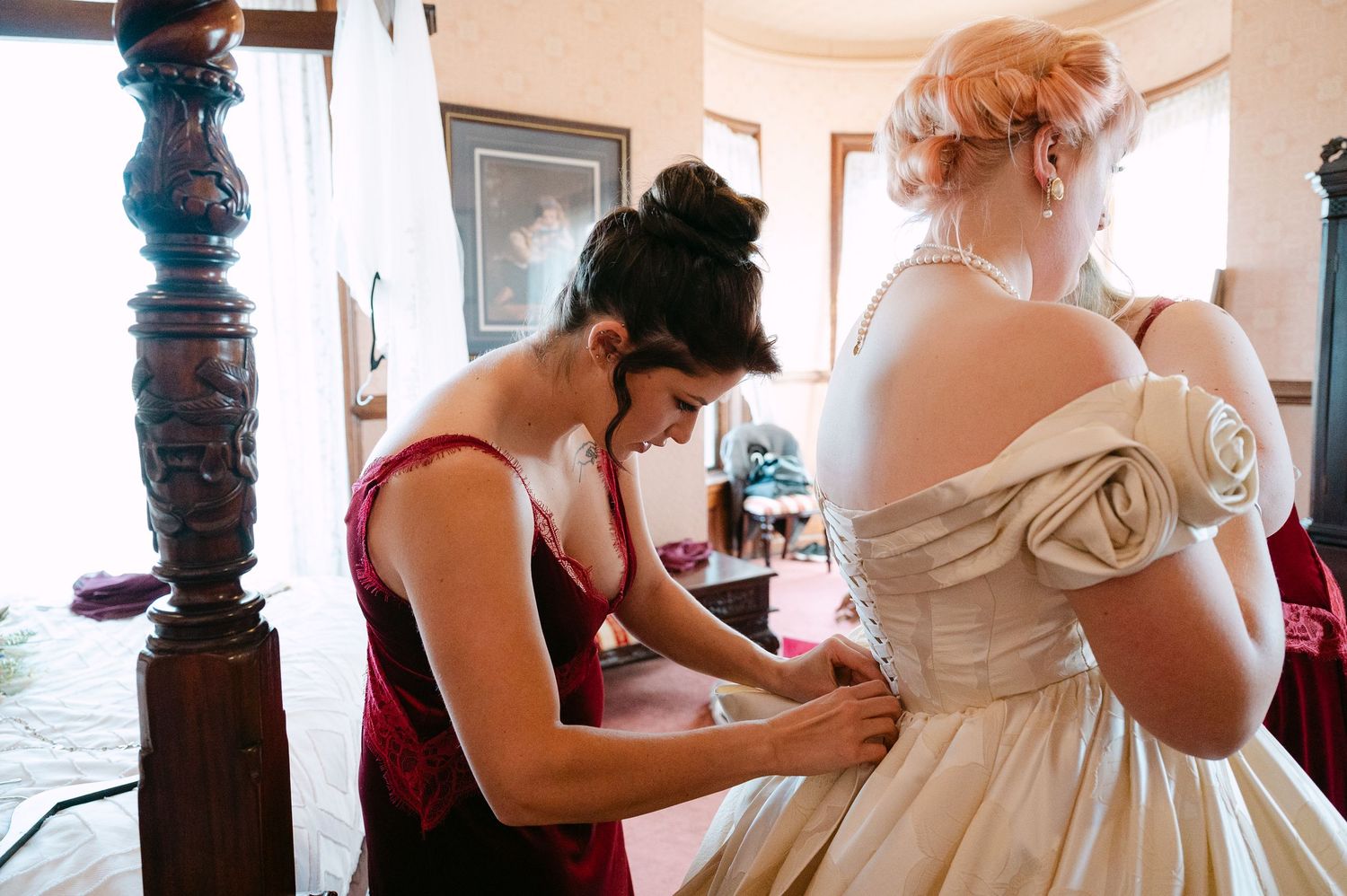 Central MN Wedding Photos | Bridesmaid in burgundy dress fastening buttons on bride's off-shoulder champagne satin wedding gown.