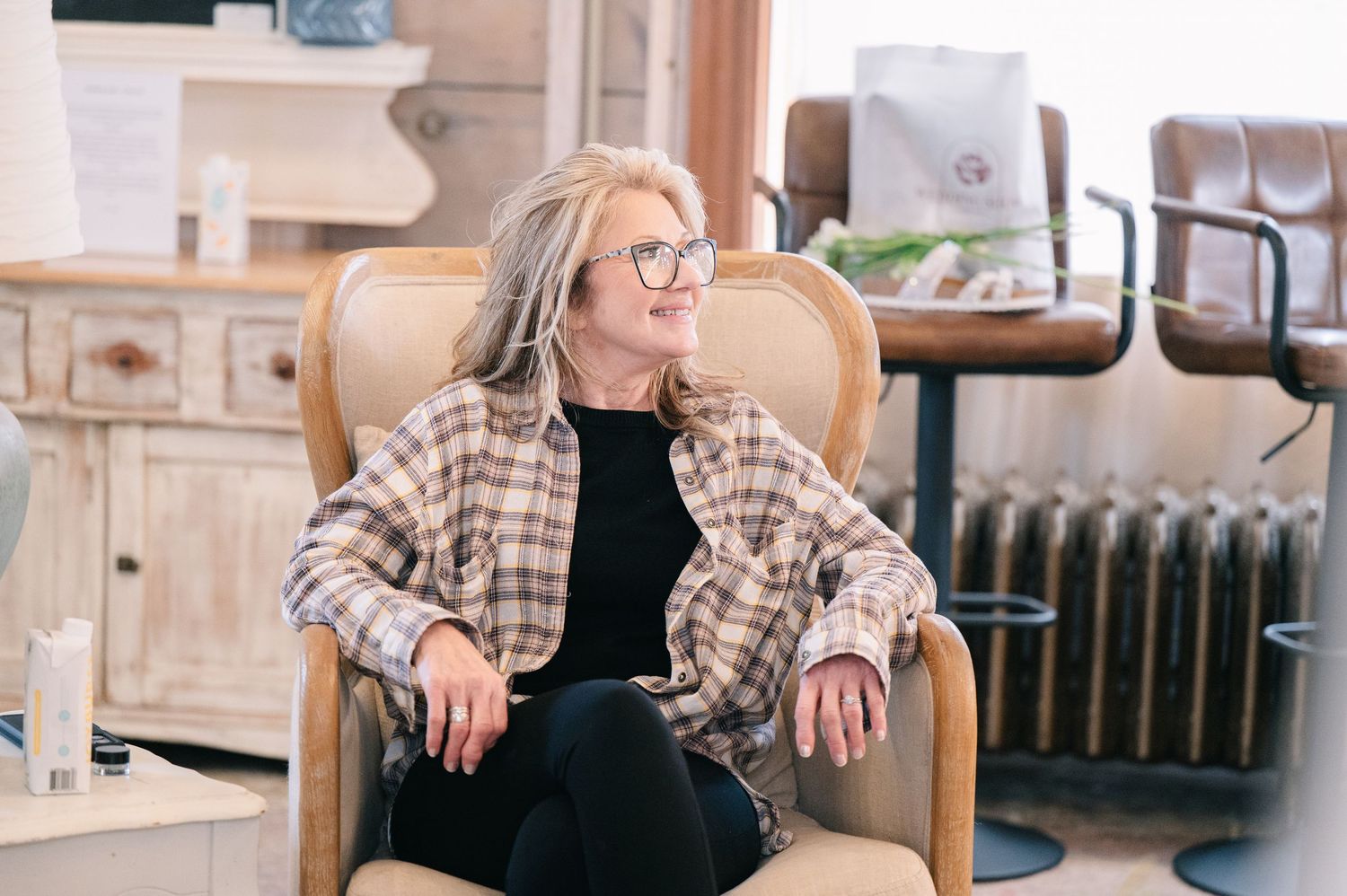 Woman with gray hair and glasses sitting relaxed in a beige armchair inside a bright, stylish interior space.