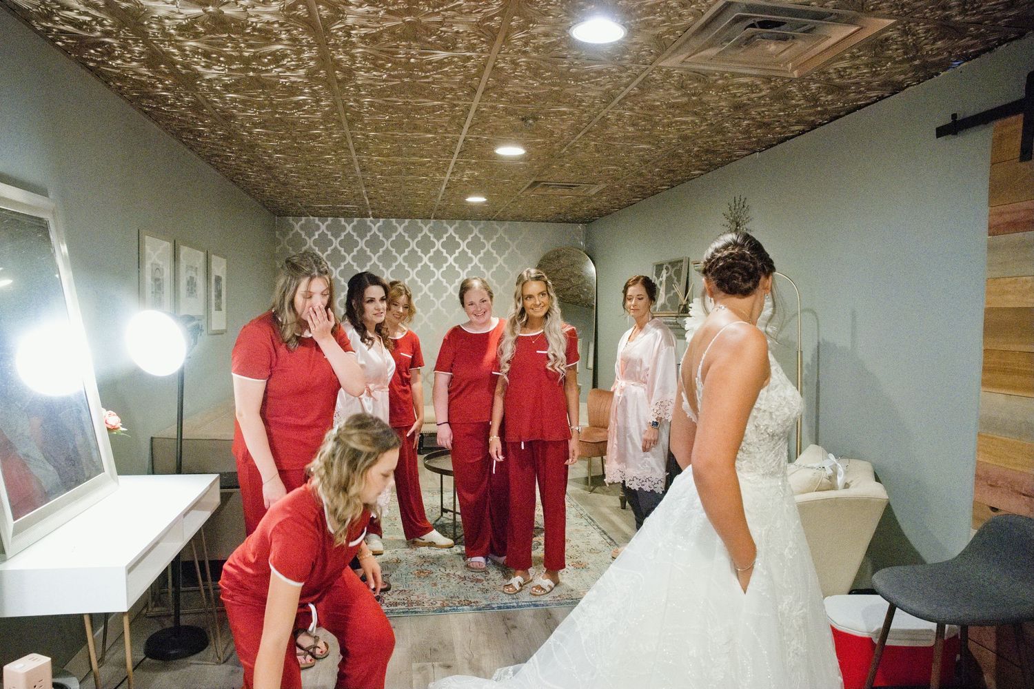 Bridesmaids in red outfits admire bride in white wedding gown in bridal suite.