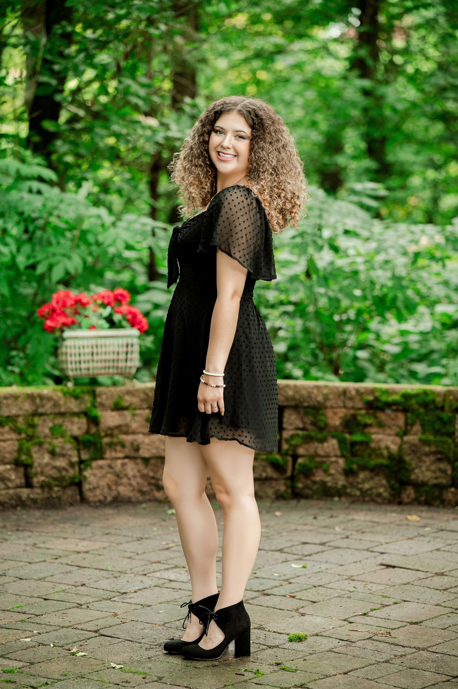 A person in a black lace dress poses on a stone patio surrounded by lush greenery and red flowers.