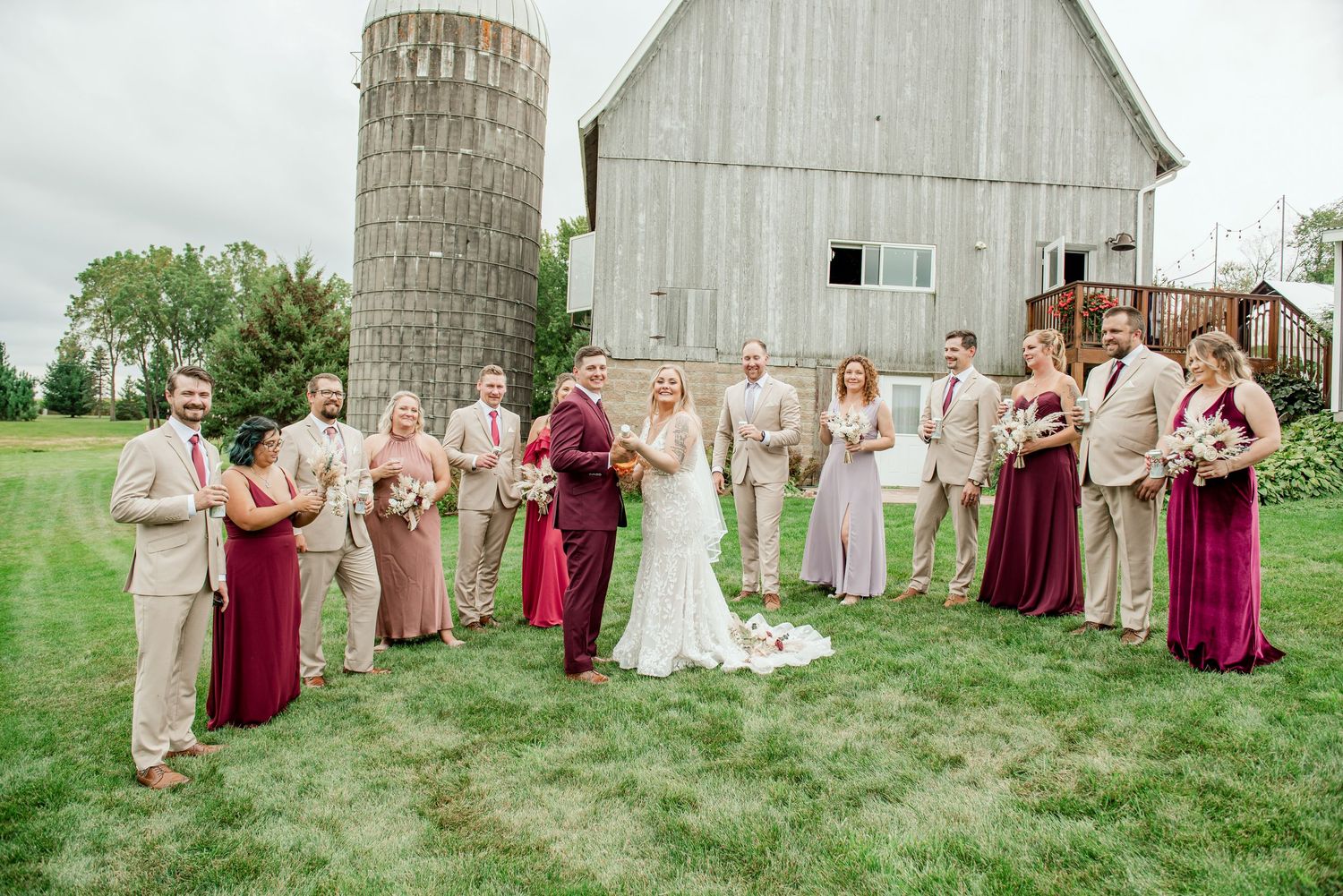 Wedding party poses in front of a rustic barn and silo wearing burgundy and tan attire in a country setting.