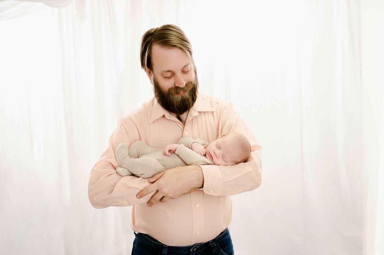 Someone wearing a peach-colored button down shirt holds a sleeping newborn baby against their chest in a bright setting.