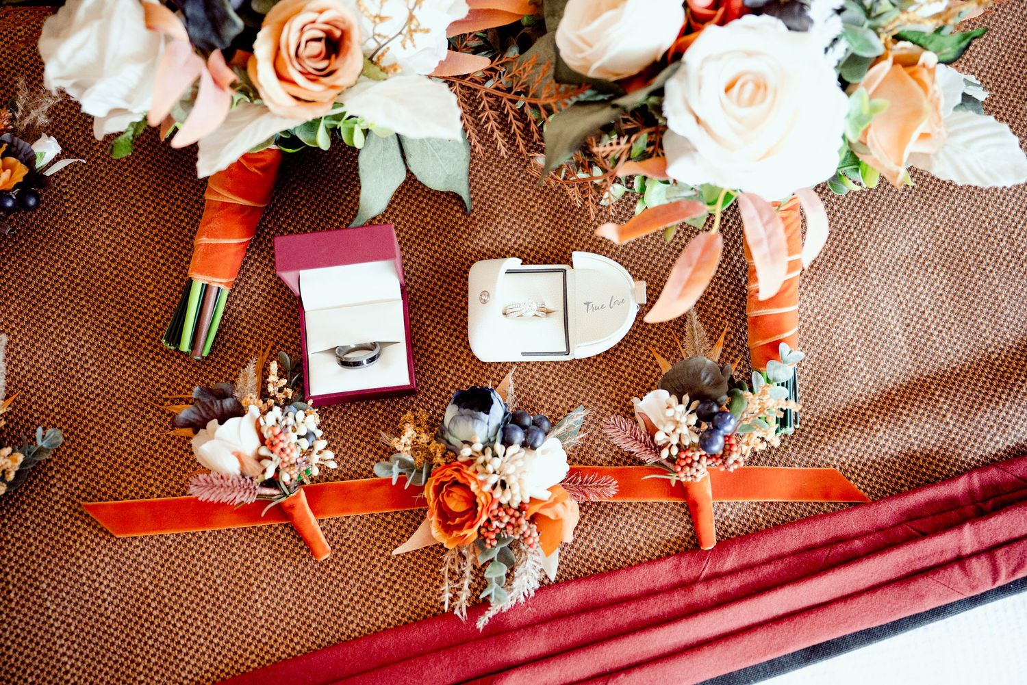 Wedding details flatlay with peach and white roses, rings, boutonnieres, and invitation on brown leather surface.