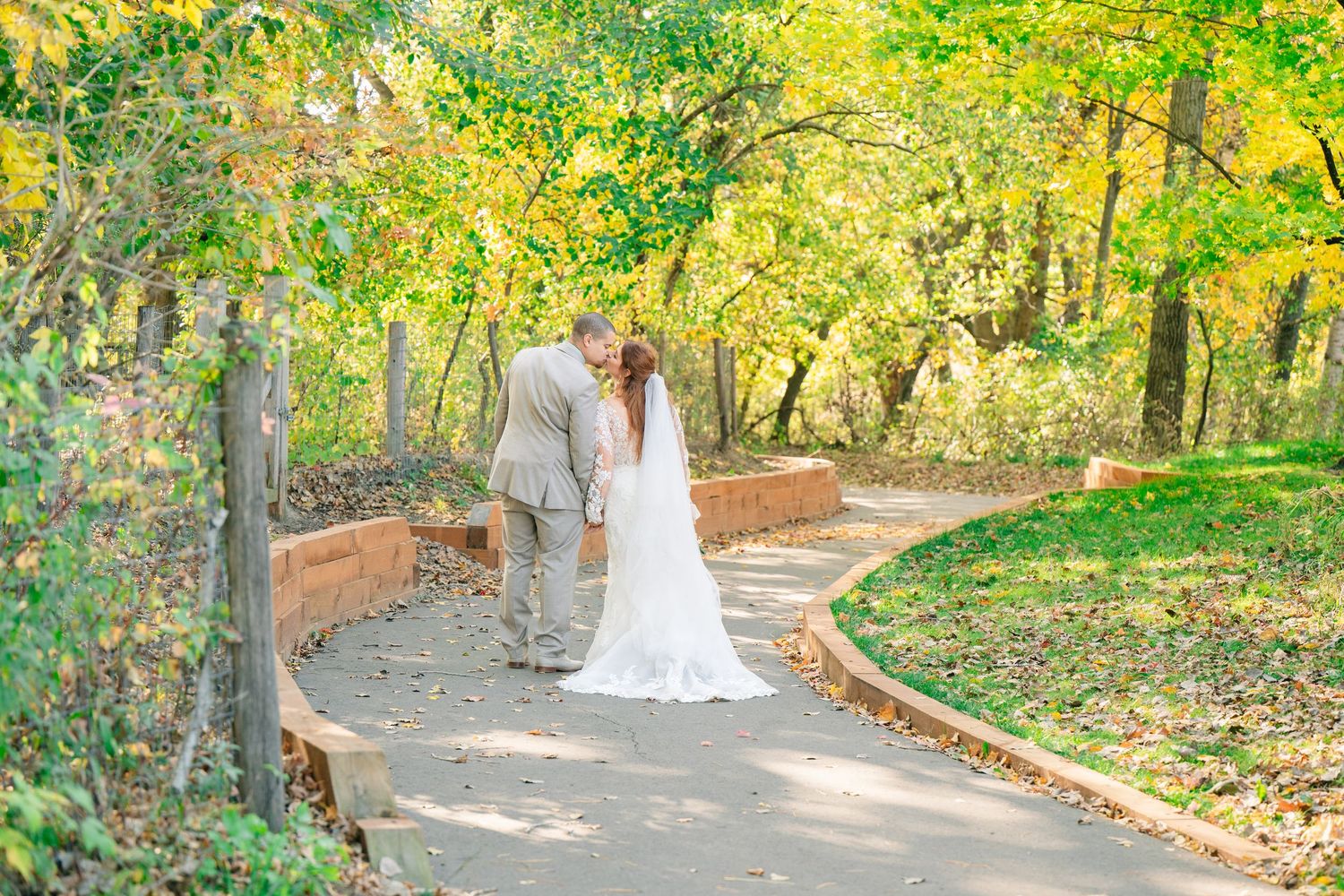 Bride and groom share a kiss on a winding garden path surrounded by lush autumn trees.