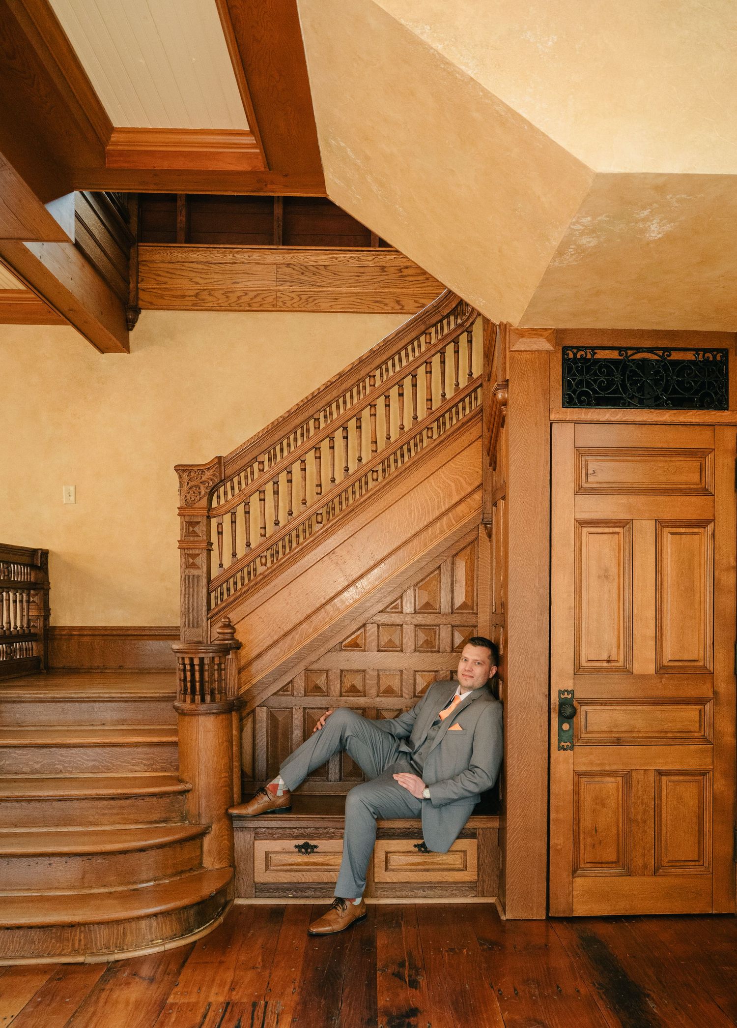 Man in gray suit sitting under ornate wooden staircase in elegant Victorian-style home with rich hardwood floors.