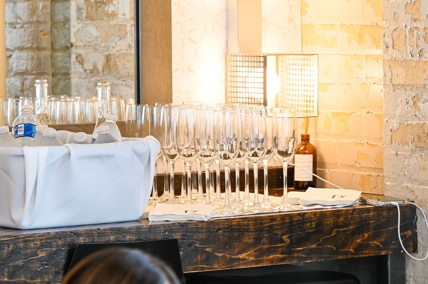 Empty wine glasses lined up on rustic wooden table with white linens and stone wall background in warm lighting.