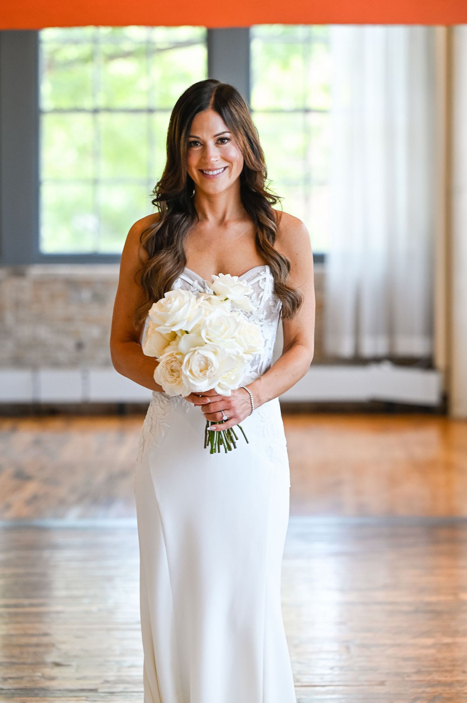 Bride in white dress holds elegant bouquet of white roses against wooden floor backdrop.