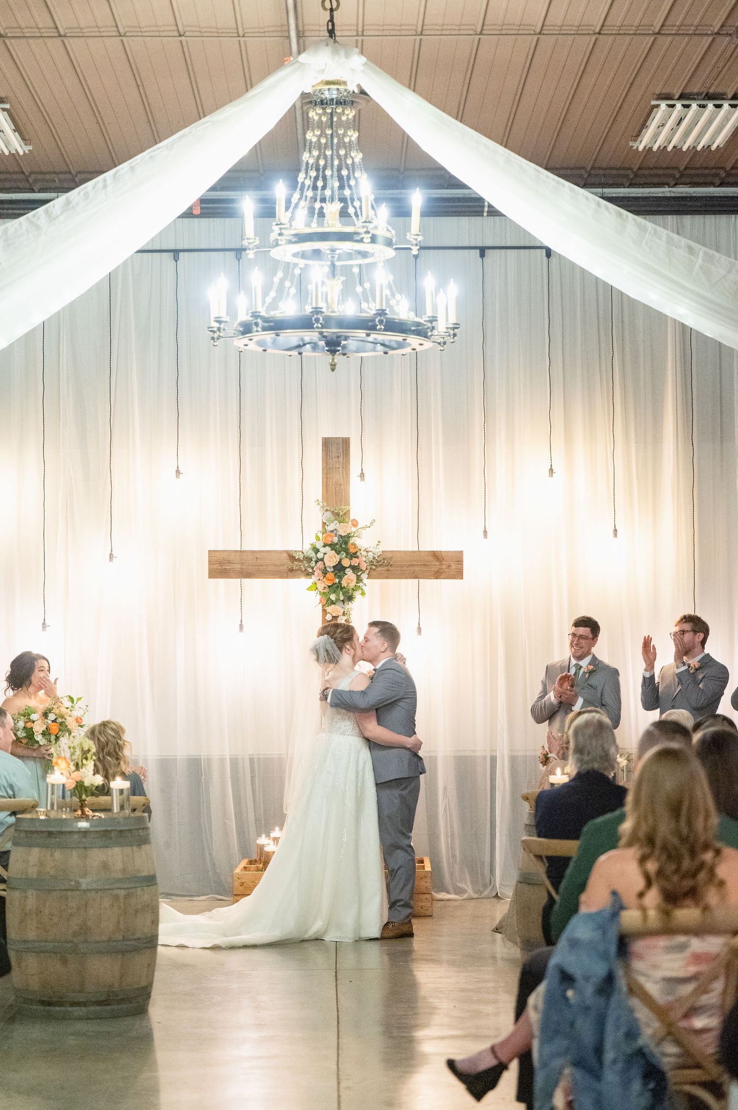 Newlyweds share first kiss under chandelier at elegant barn wedding ceremony.