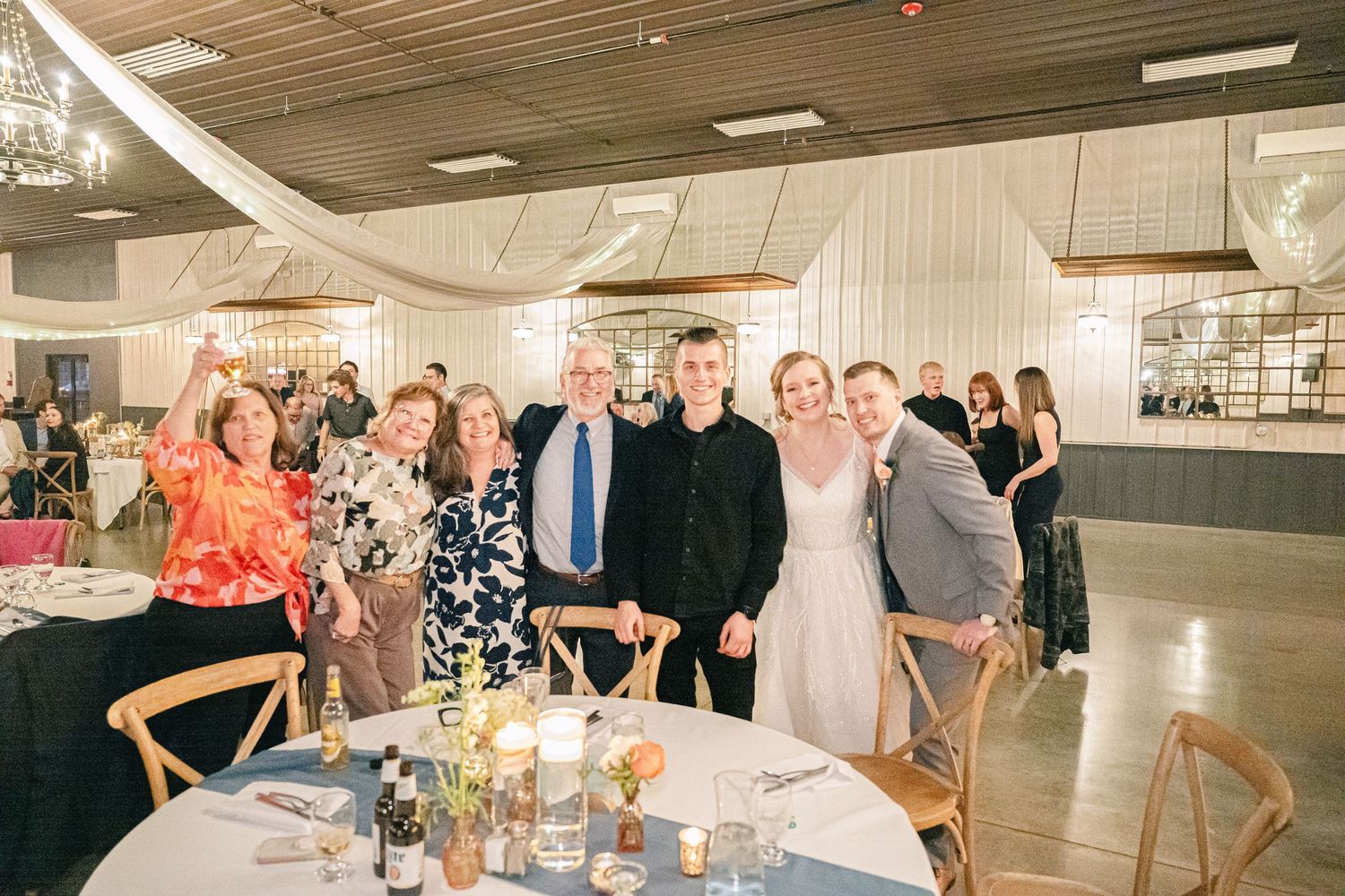 Bride and groom pose with family members at decorated reception table in elegant wedding venue.