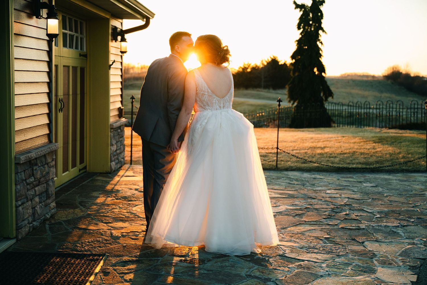 Bride and groom kissing at sunset outside a rustic wedding venue.
