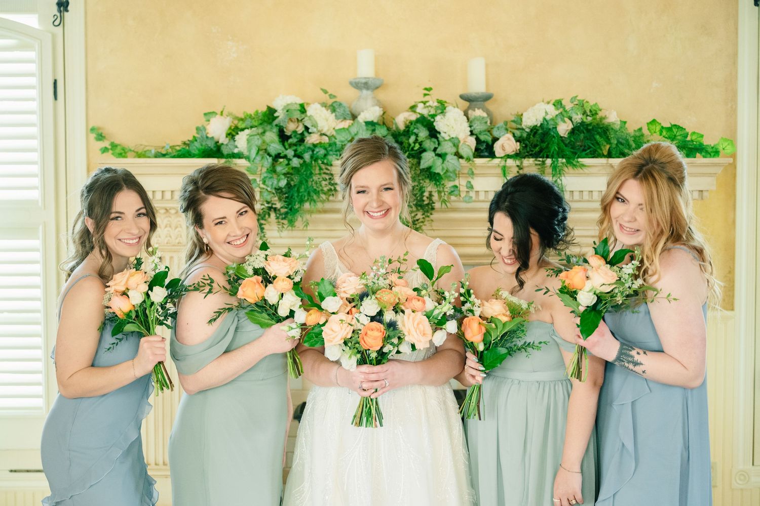 Bride and four bridesmaids in sage green dresses holding peach and white bouquets in front of floral fireplace.