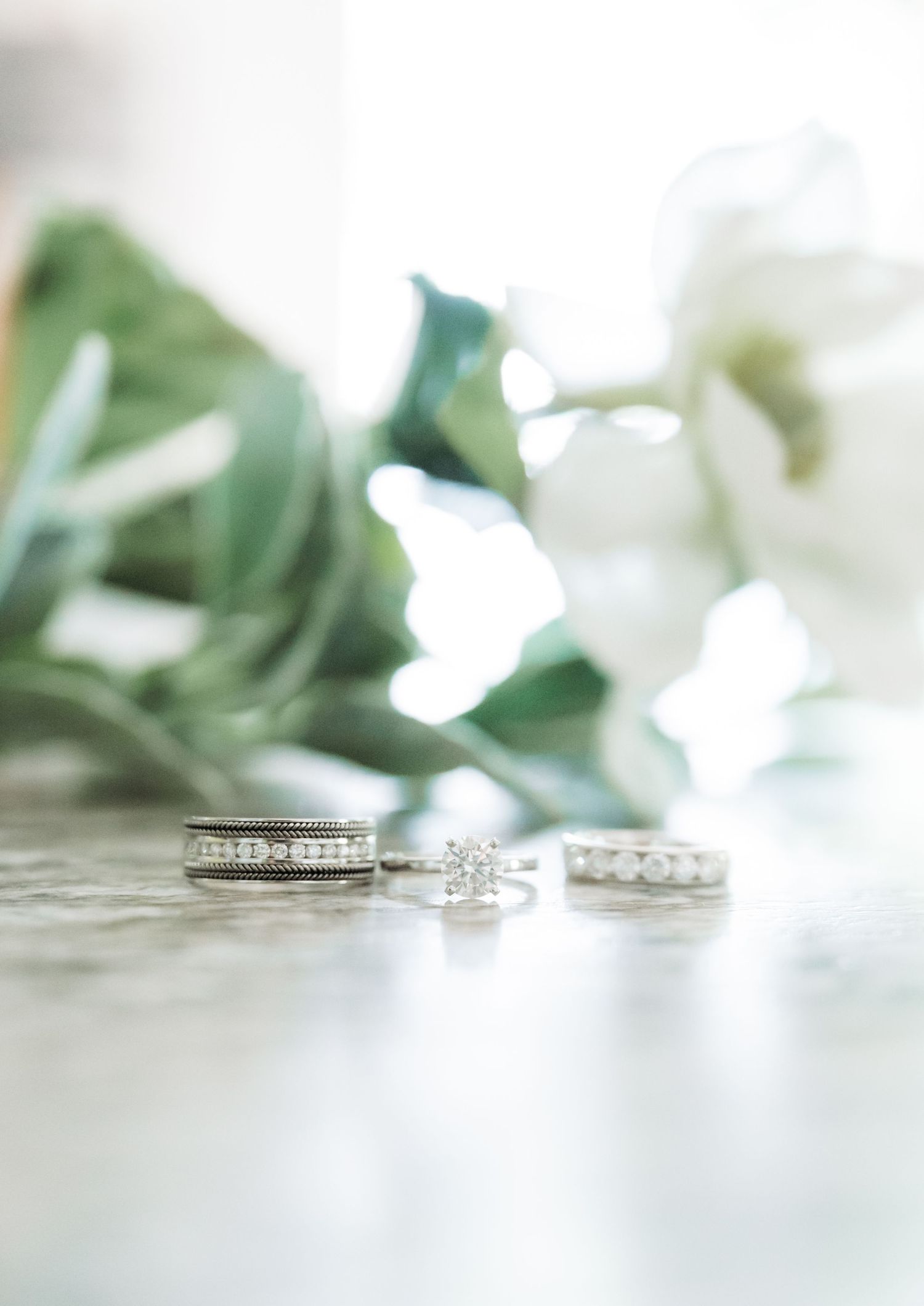Three diamond wedding rings arranged on a marble surface with soft white flowers and greenery in the background.