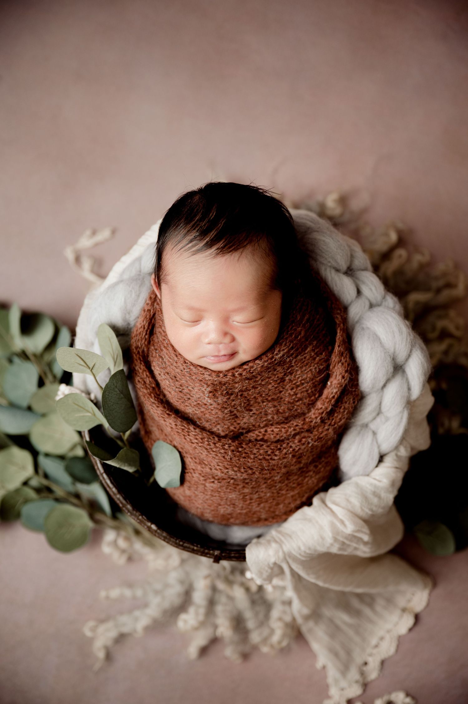 St Cloud newborn Photos of Newborn baby wrapped in brown knit swaddle resting in a wooden bowl with eucalyptus greenery.