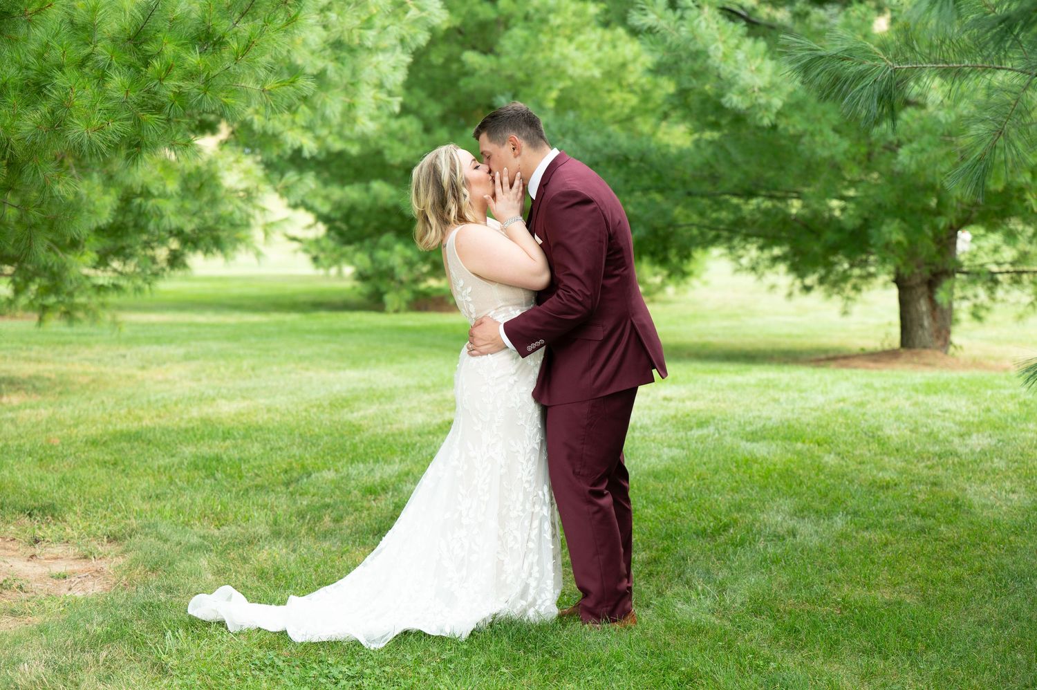 Central MN Wedding Photography - Bride and groom share a tender kiss in a sunlit green park, bride in lace gown, groom in burgundy.