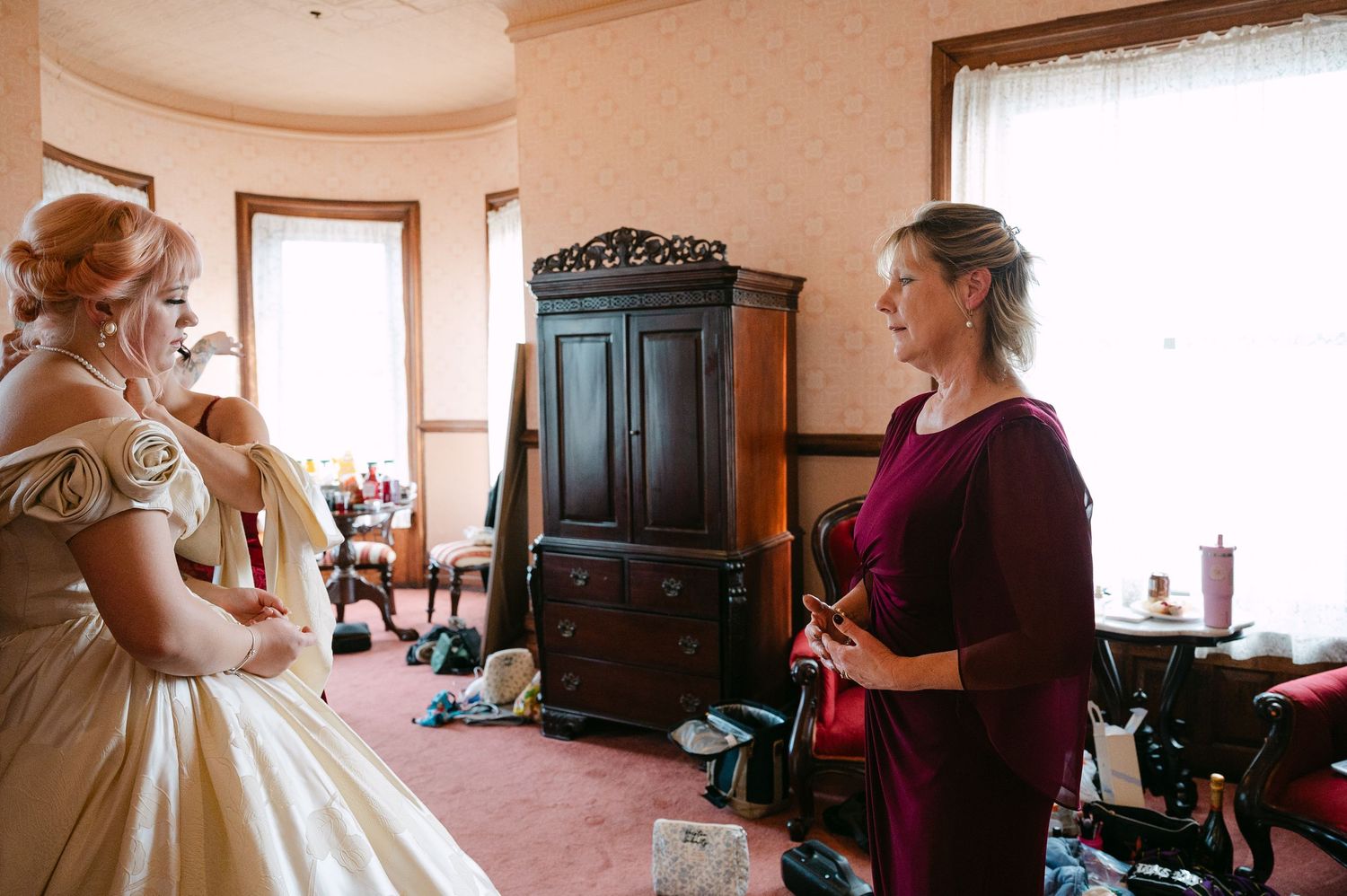 Central MN Wedding Photos | Bride in golden wedding gown getting ready with bridesmaid in burgundy dress in vintage decorated room.