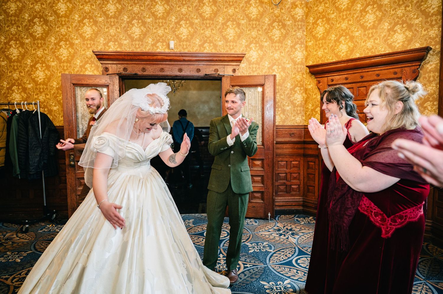 Central MN Wedding | Bride in vintage white gown twirls while groom in green suit and bridesmaid in burgundy velvet applaud in ornate room.